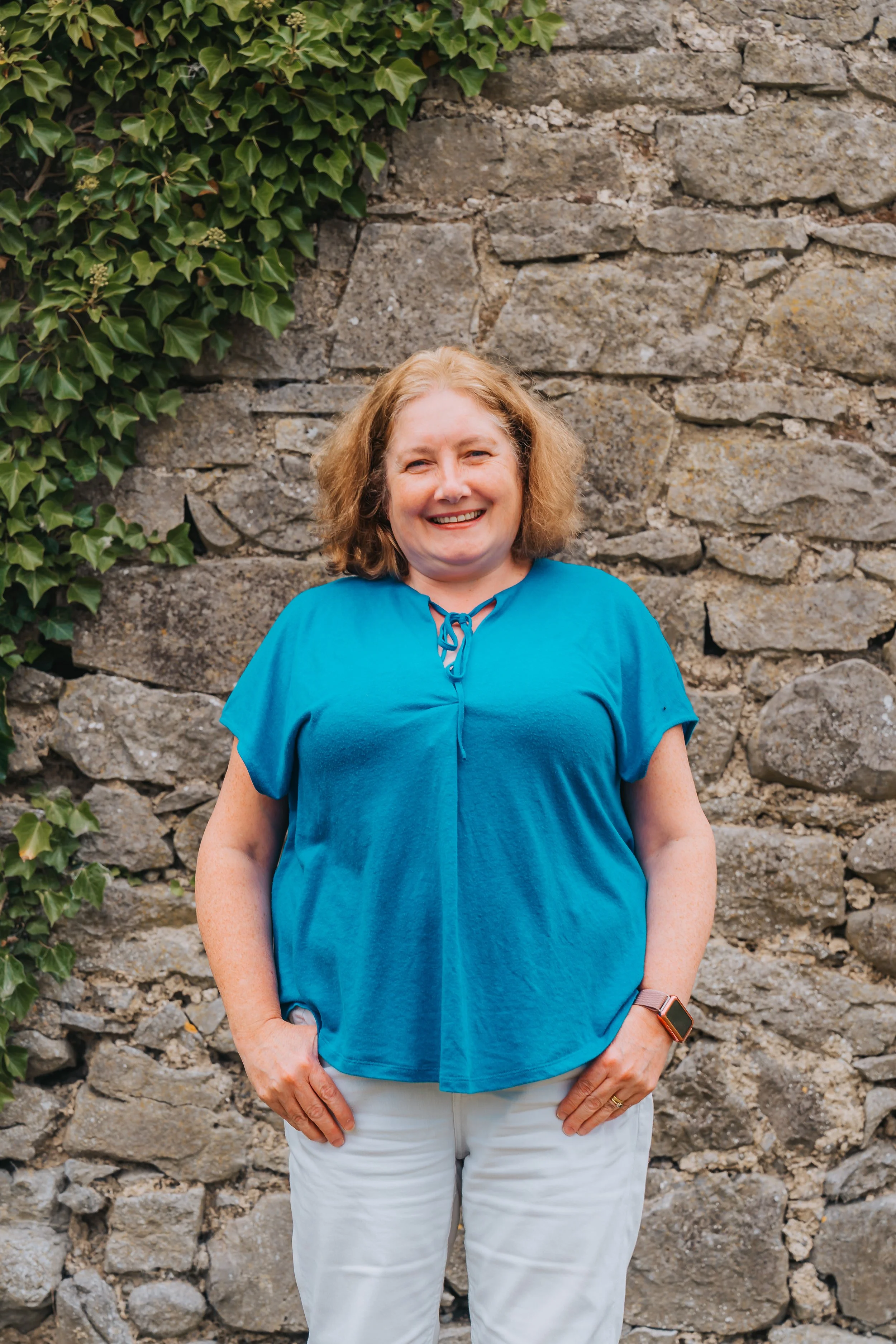 A woman with curly, shoulder-length blonde hair smiling and standing against a stone wall with green ivy leaves on the upper left. She is wearing a bright blue top with a tie at the front, light-colored pants, and a smartwatch on her left wrist.