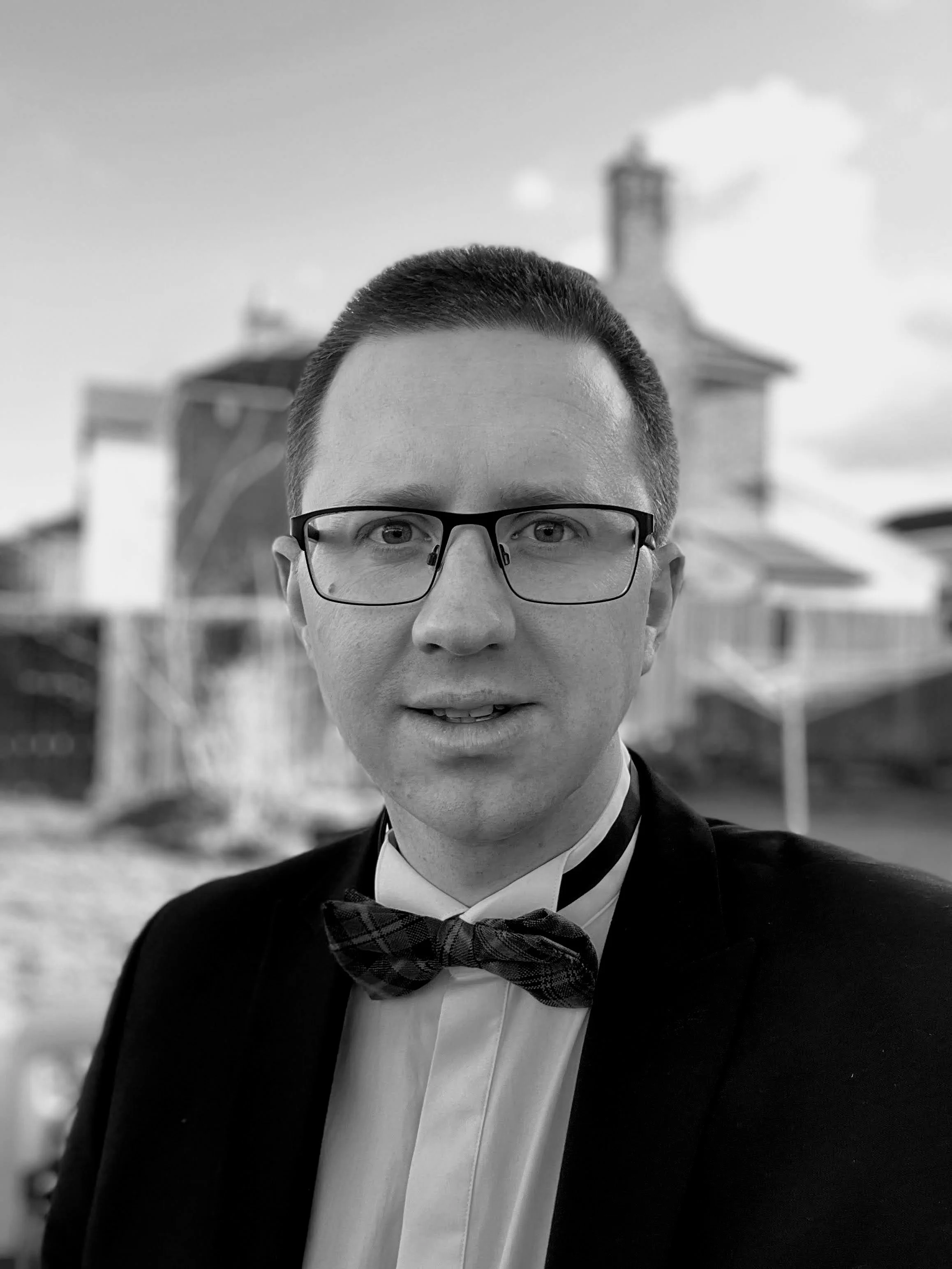 A man wearing glasses, a tuxedo, and a bow tie, smiling outdoors with a house and chimney in the background.