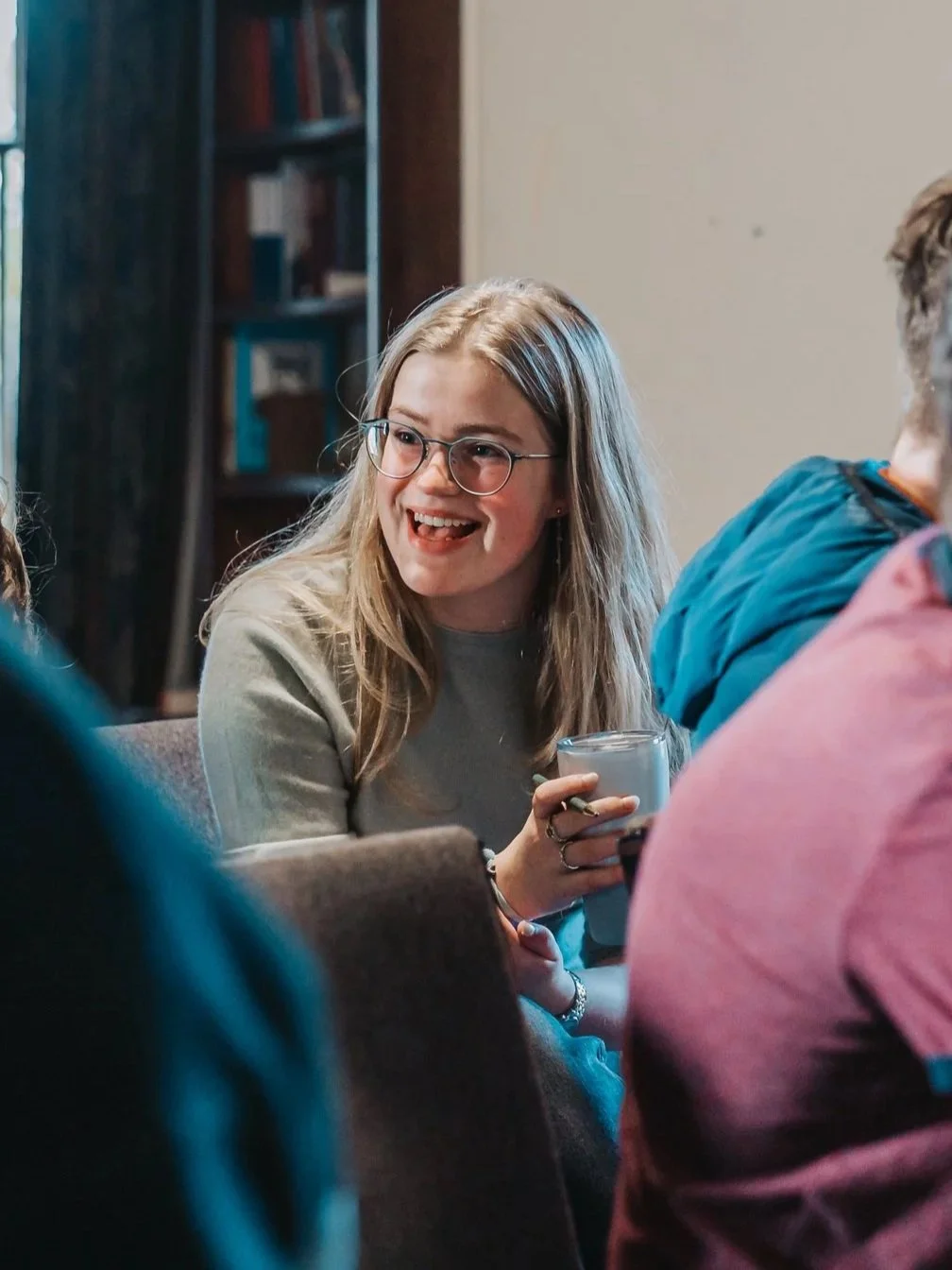 A young woman with glasses and blonde hair sitting in a room, smiling and holding a glass of water, engaged in conversation.
