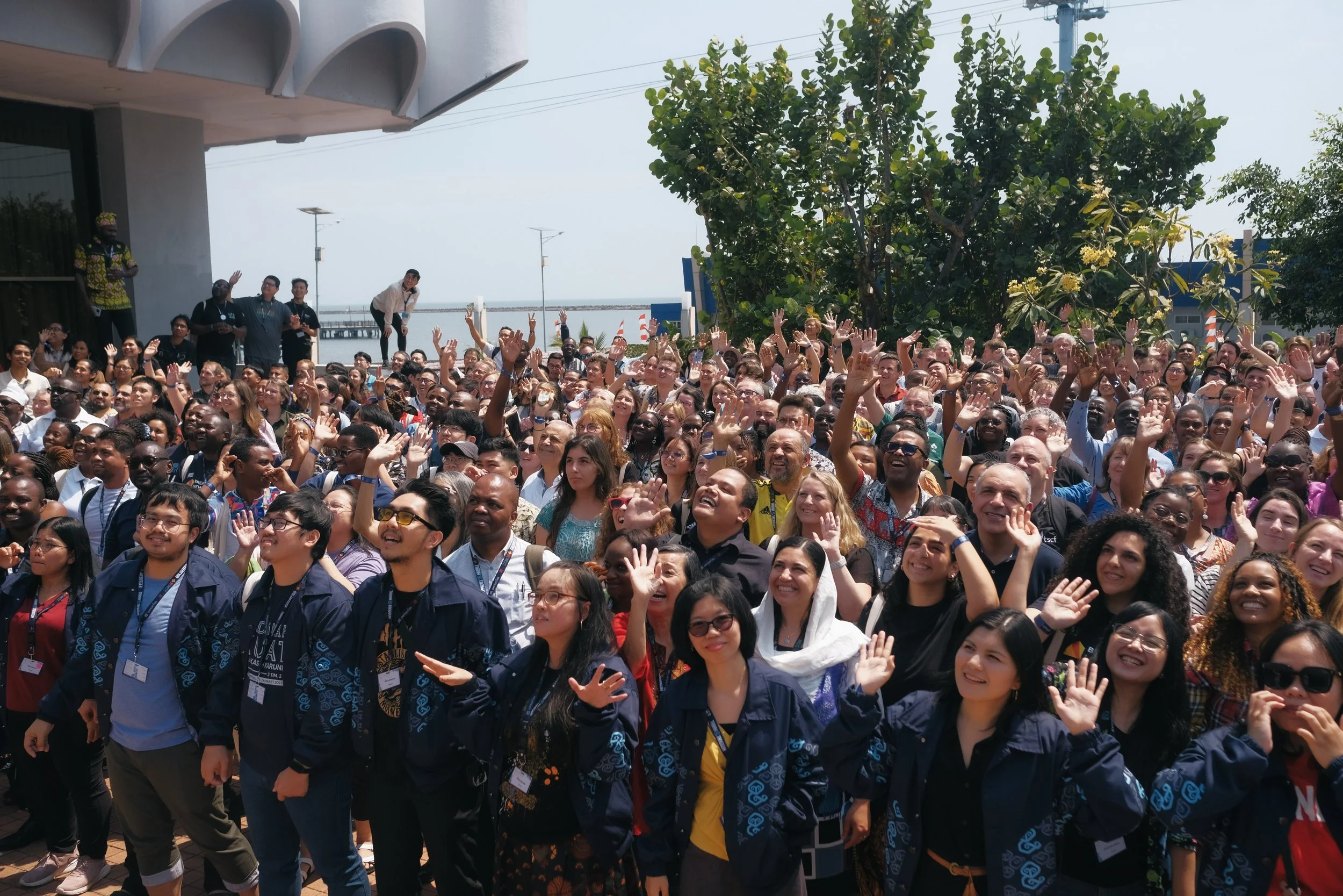A large outdoor gathering of diverse people, many with hands raised and smiling, with trees and a pier in the background.