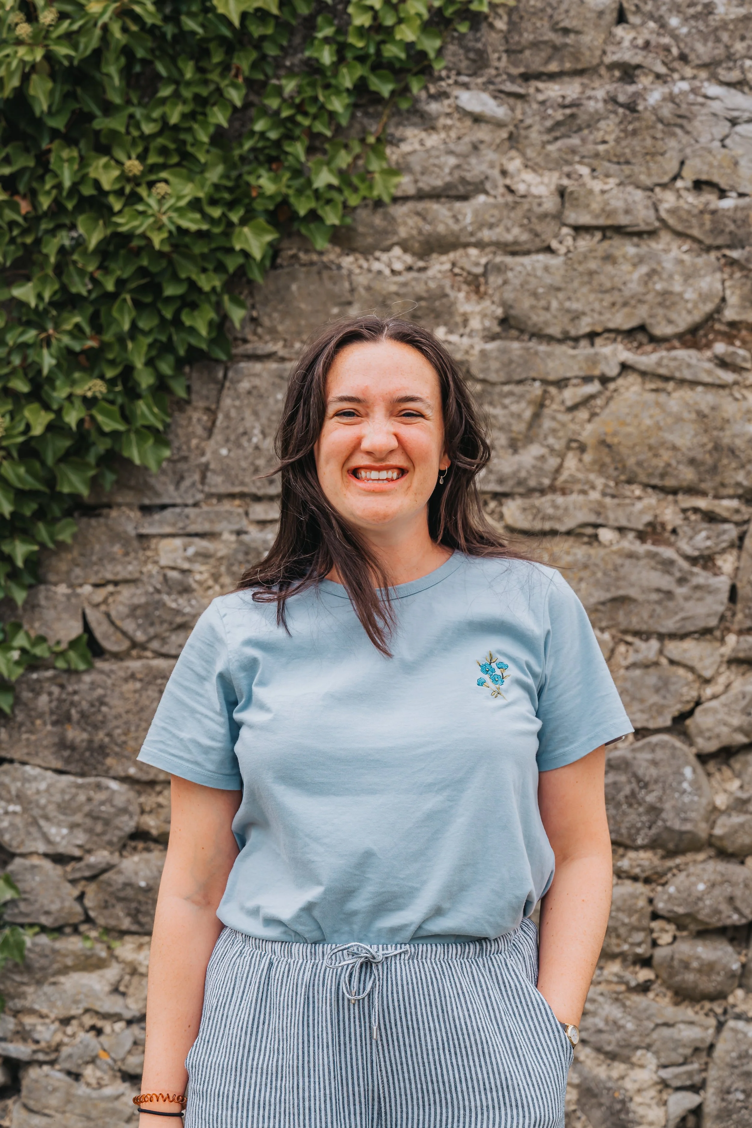 A young woman with long brown hair smiling and wearing a light blue T-shirt with a small embroidered floral design, standing in front of a stone wall with green ivy plants growing on it.
