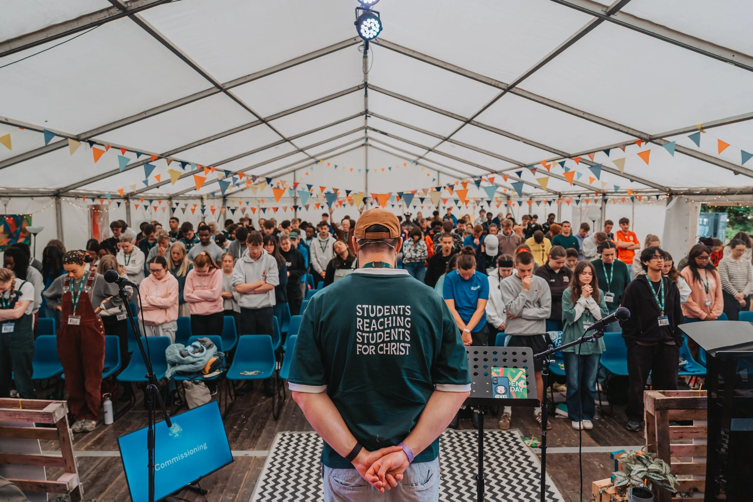A large group of people gathered inside a tent for an event, with a man in the foreground wearing a shirt that reads "Students reaching students for Christ." The crowd appears to be listening to a speaker or presentation, with colorful triangular banners hanging from the ceiling.