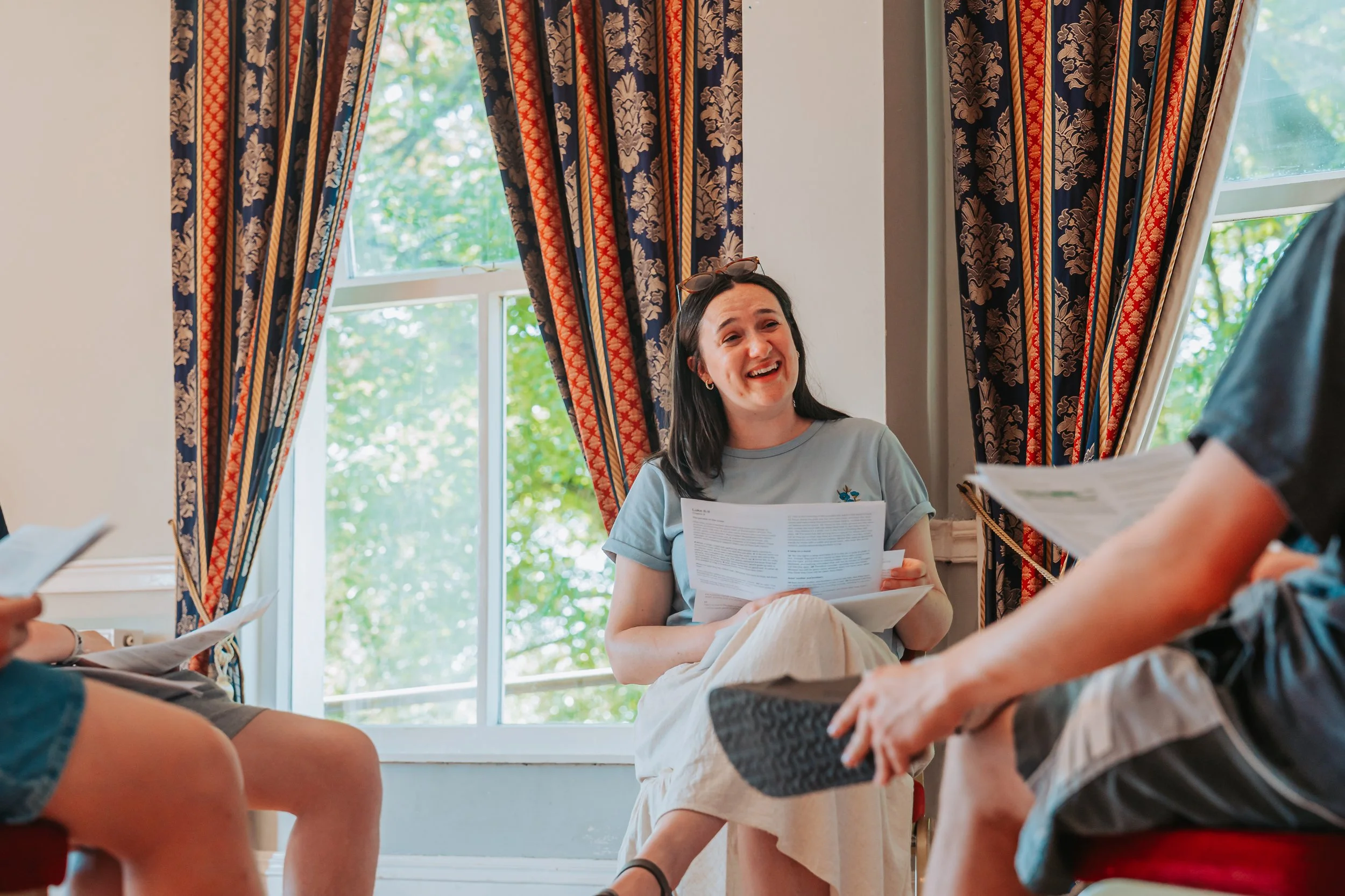 A young woman with dark hair, wearing a gray shirt, sitting by a window with floral curtains, smiling and holding papers while talking to several people whose legs are visible in the foreground.