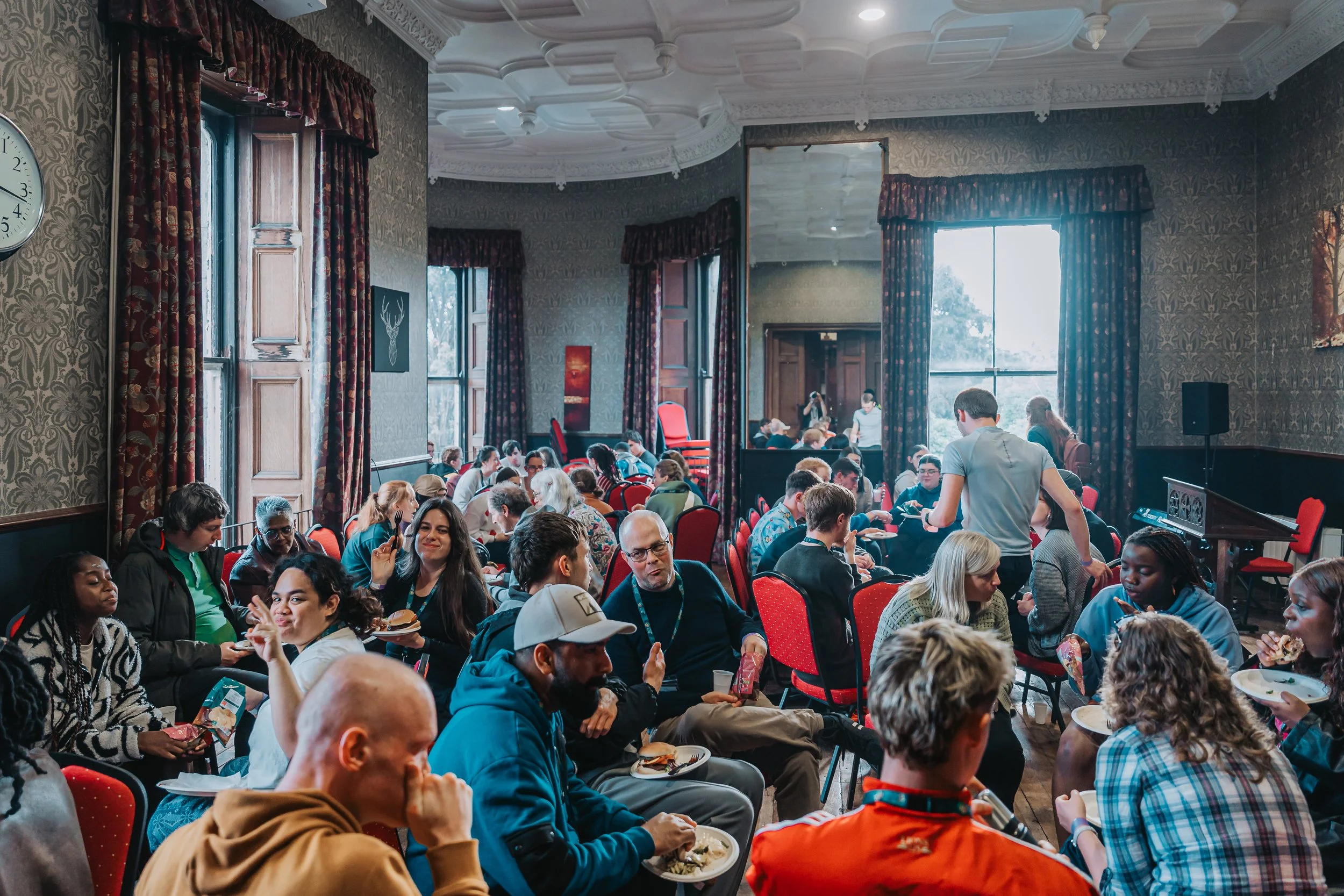A large indoor gathering or conference with many people seated around tables, eating, talking, and mingling. The room features high ceilings, ornate crown molding, tall windows with floral patterned curtains, and a large mirror on the wall.