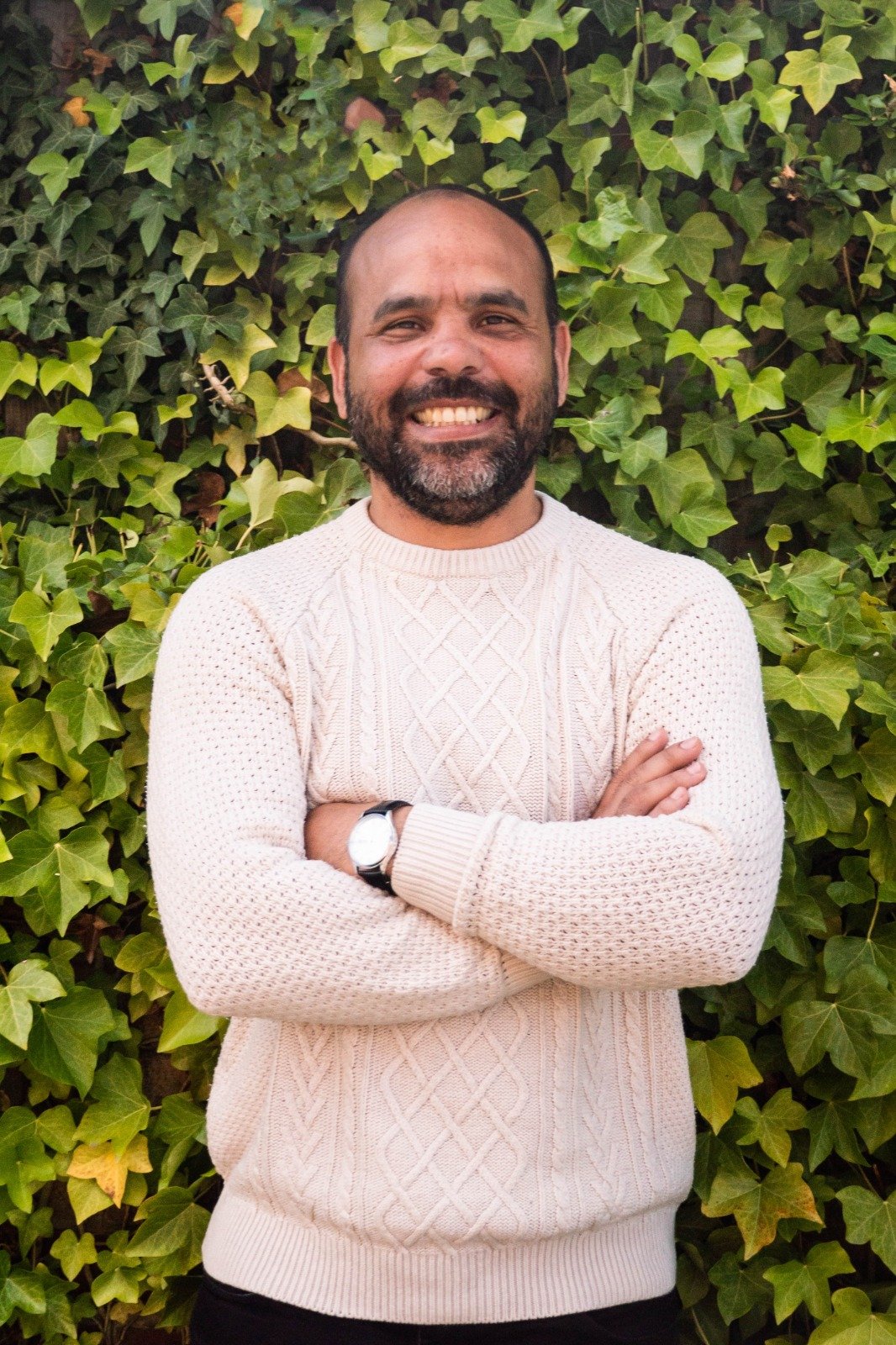 A smiling man with a beard and short hair, wearing a cream-colored cable-knit sweater, arms crossed, standing in front of a lush green ivy-covered wall.