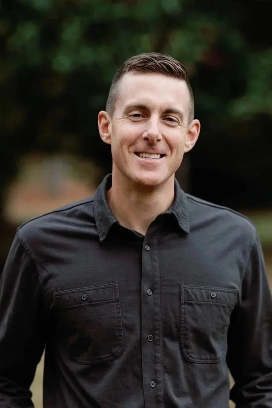 A smiling young man with short brown hair, wearing a black button-up shirt, outdoors with blurred greenery in the background.