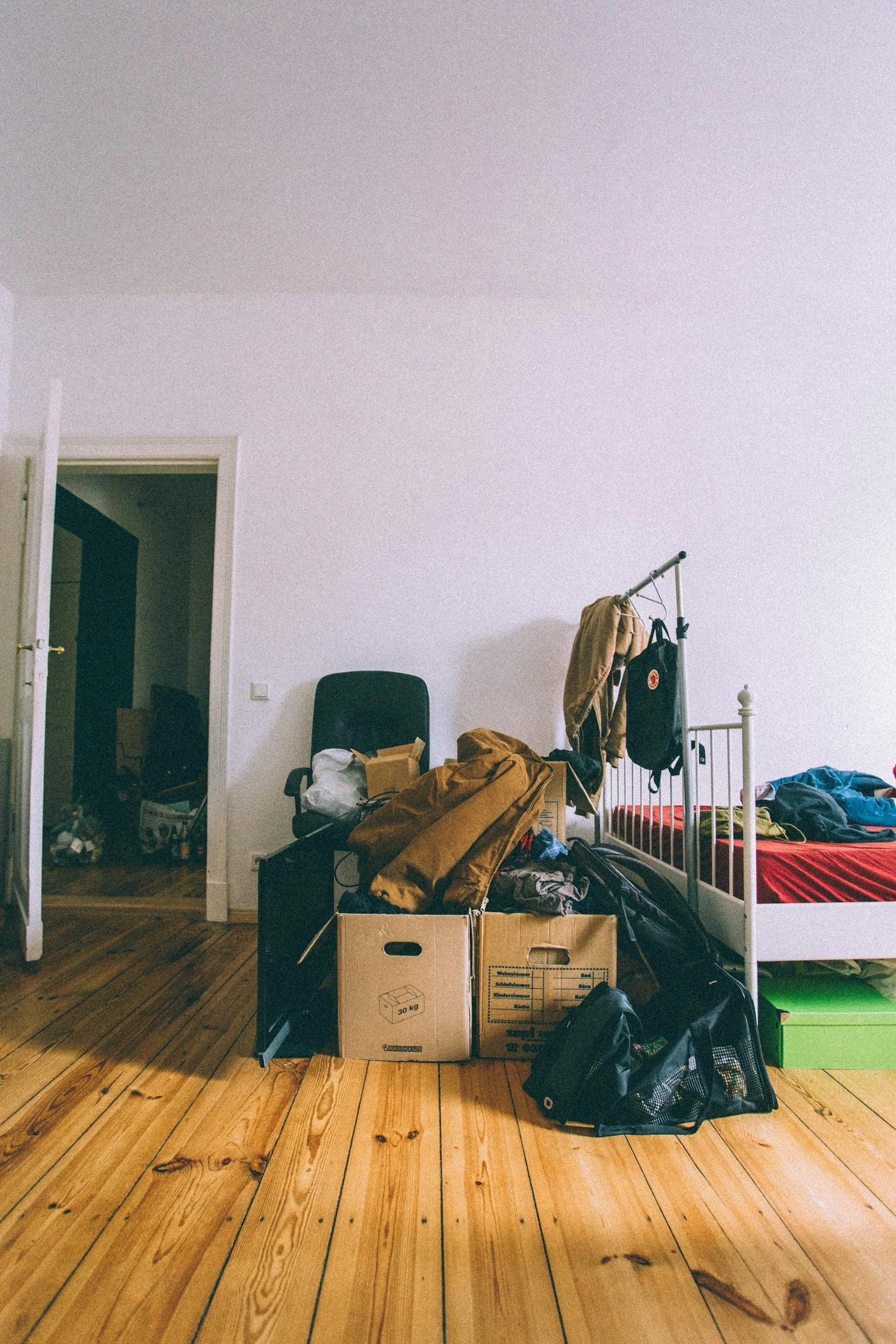 Bedroom with a white bed frame, cluttered with various bags, boxes, and clothes, and a tall white coat rack with clothes hanging on it. The room has wooden flooring and an open door leading to a dark hallway.