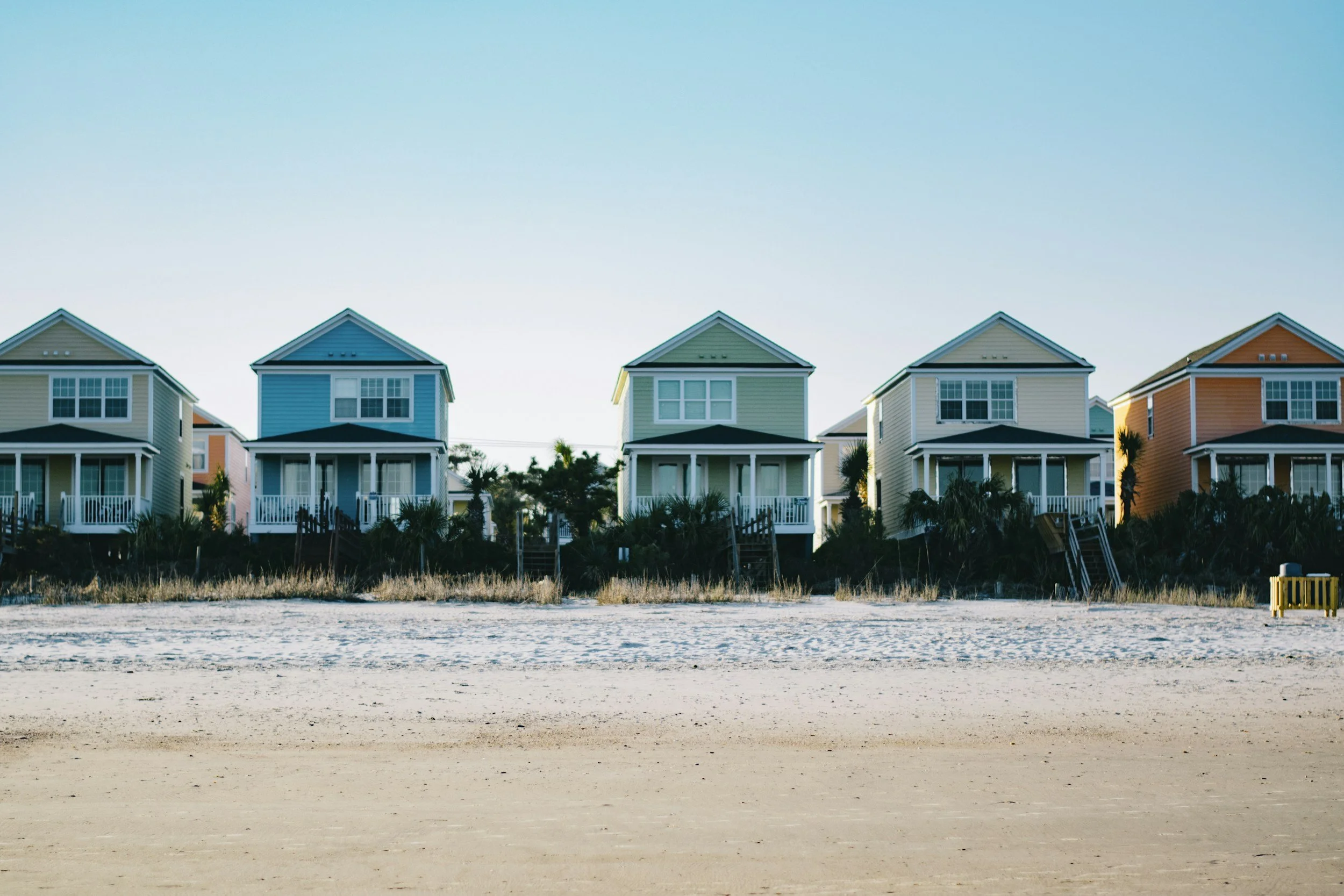 Colorful beach houses with porched entrances are lined up along a sandy shoreline with vegetation in the foreground, under a clear blue sky.