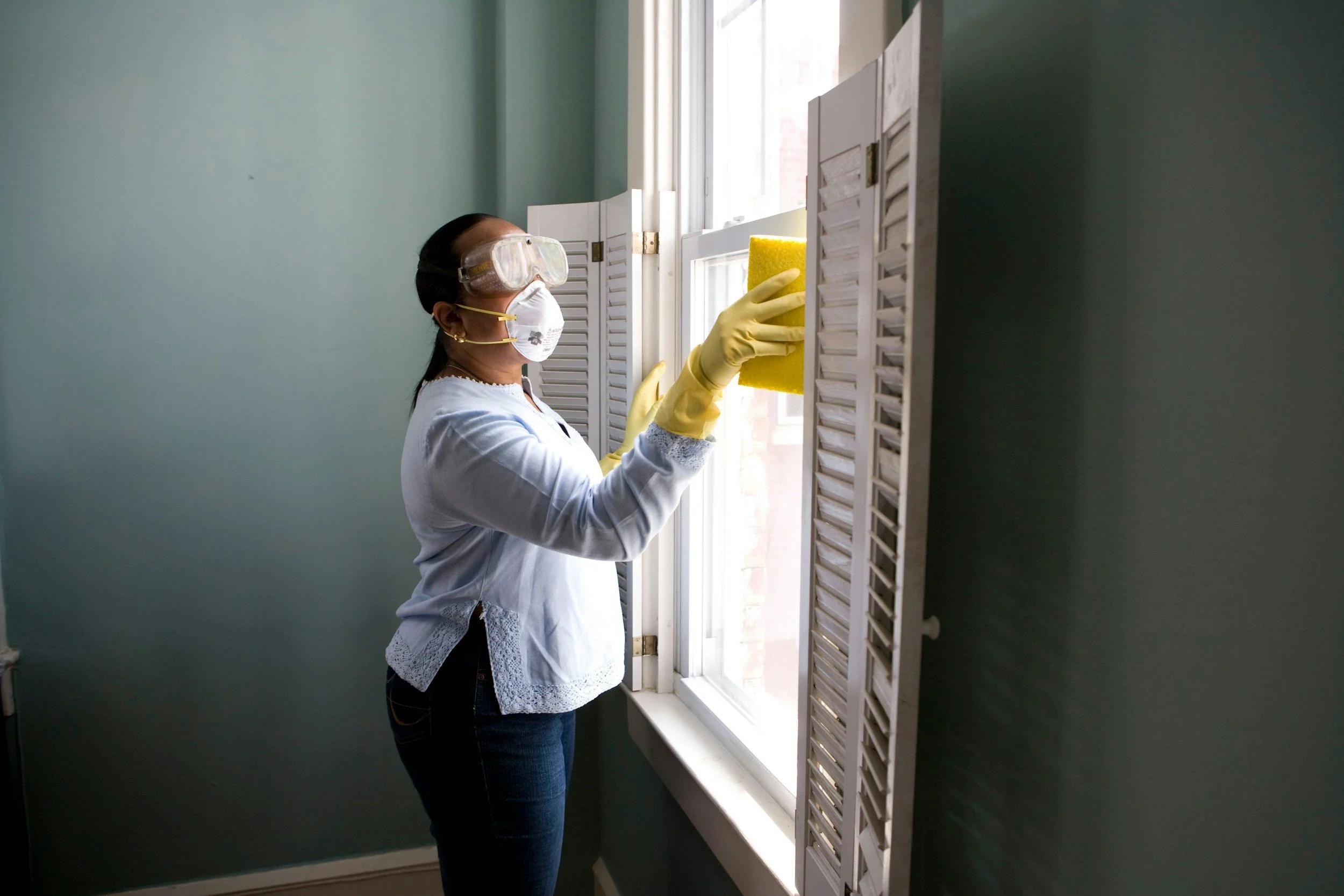 A woman cleaning a window with a yellow sponge, wearing safety goggles, a white face mask, yellow gloves, and a white blouse with lace details.
