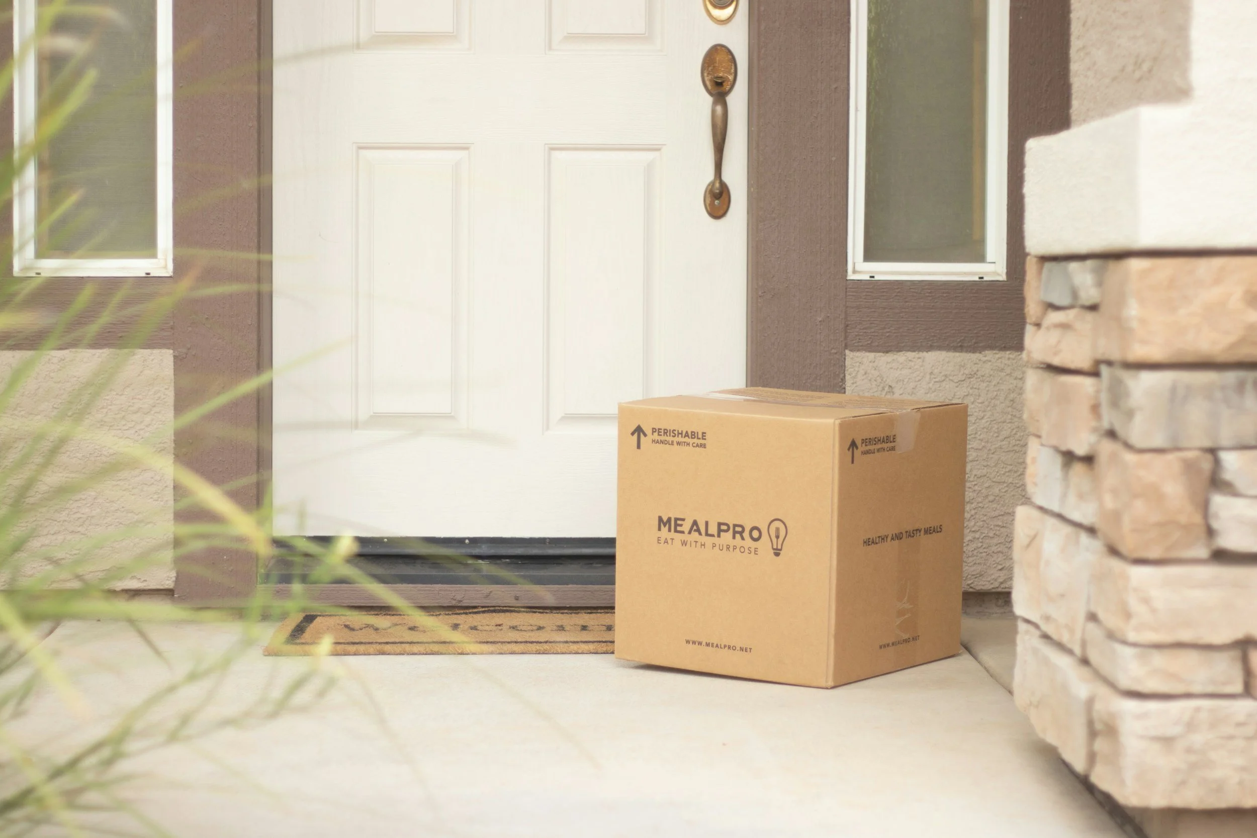 A closed white front door with a metal handle and a cardboard delivery box on the doorstep. The box has the MealPro logo and the words "Eat with Purpose" printed on it. There is a brick wall on the right and a house window on each side of the door.