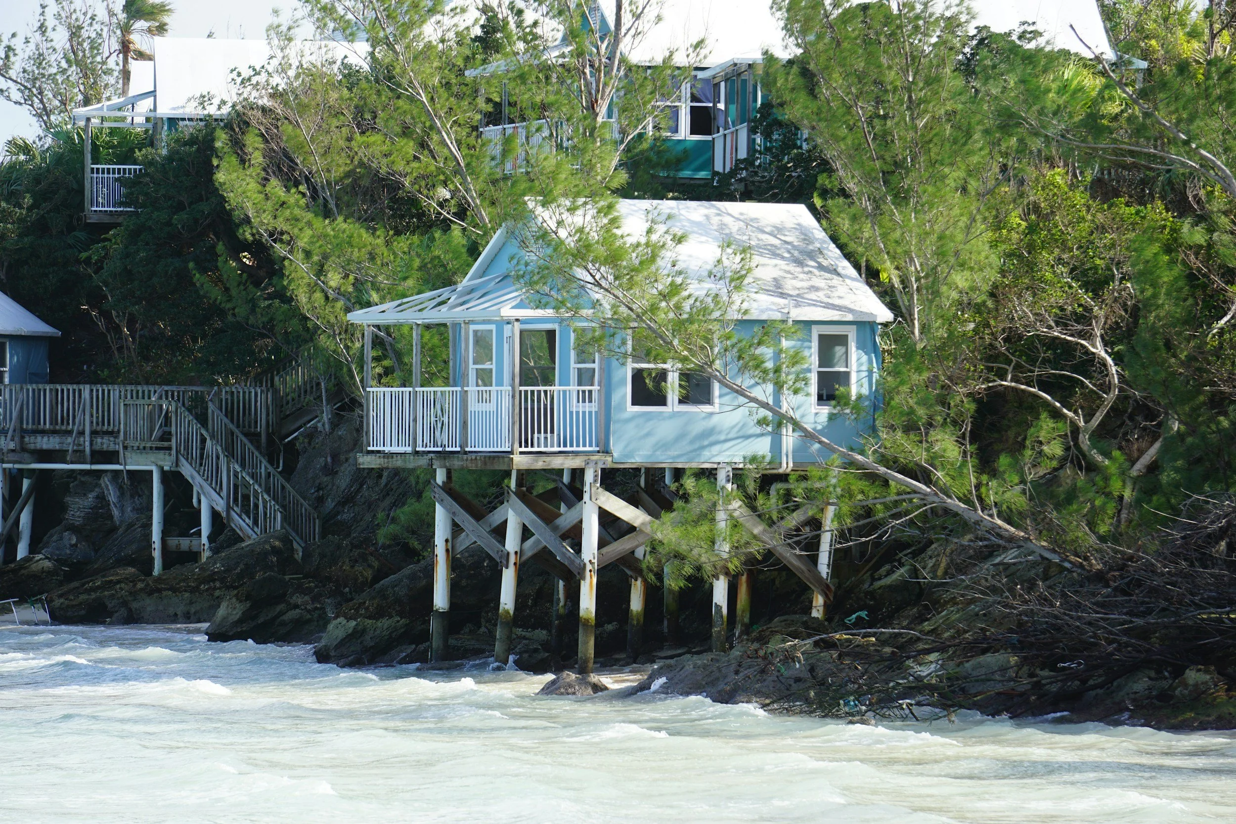 A house built on stilts on the shoreline surrounded by trees, with the ocean in the foreground and rocks beneath the house.