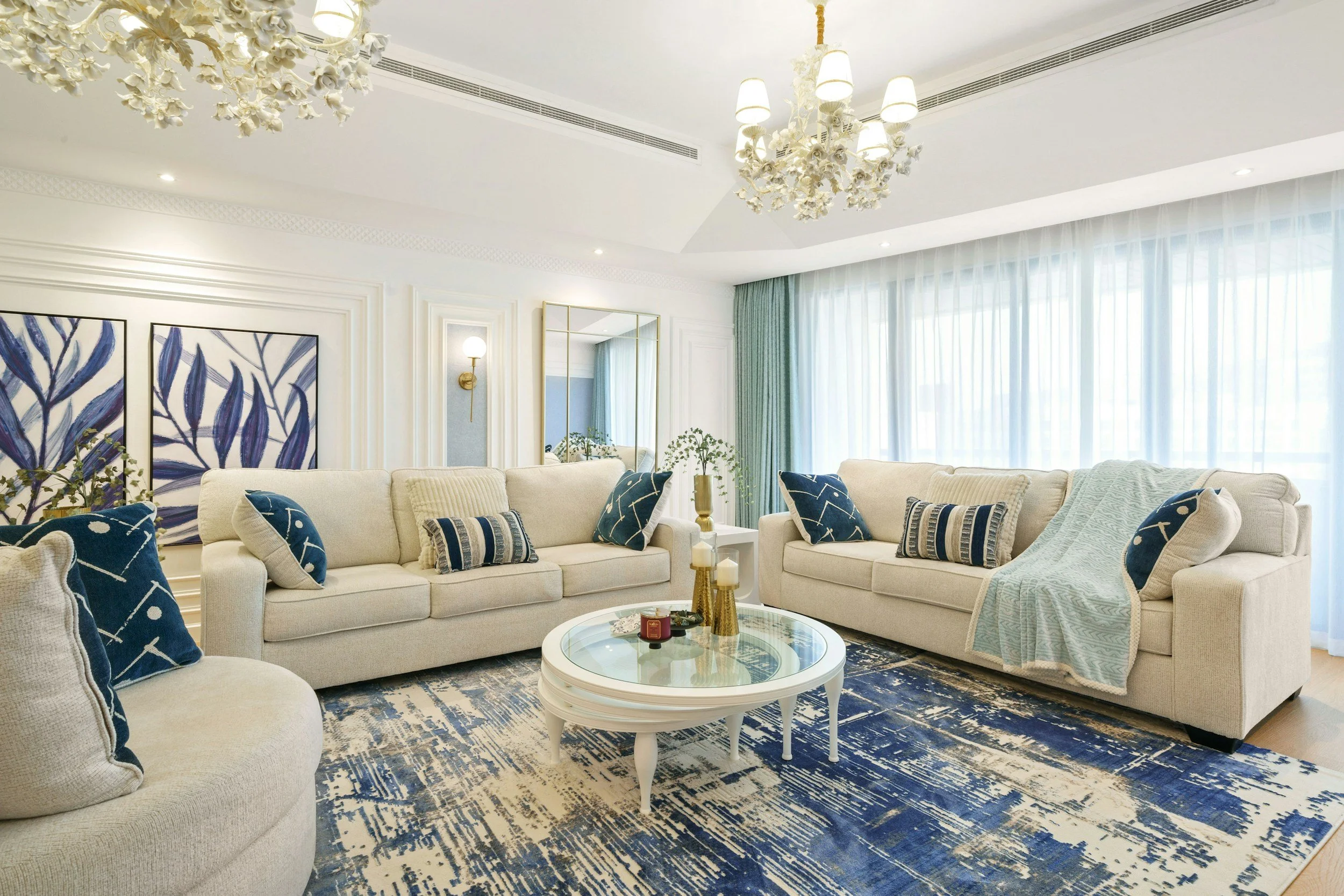 A living room with a cream-colored sofa set decorated with blue and white patterned pillows, a round glass coffee table with decorative vases and candles, large windows with sheer white curtains and green drapes, artwork of blue leaves on the wall, a mirror, a wall sconce, and a floral chandelier hanging from the ceiling.