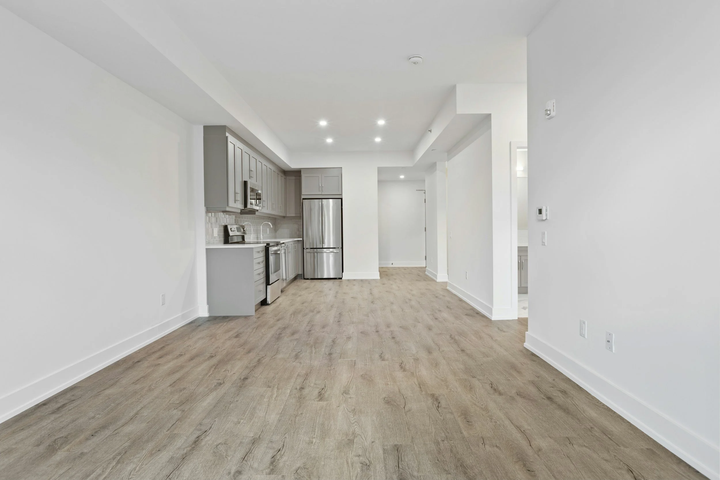 Empty modern kitchen with gray cabinets, stainless steel appliances, wooden flooring, and white walls.