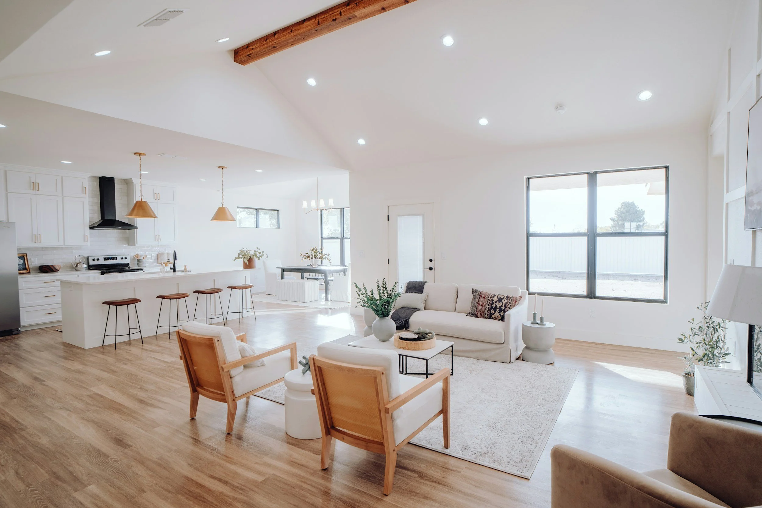 A spacious, modern open-plan living area with white walls and large windows, featuring a kitchen with white cabinets, a black range hood, and a central island with bar stools. The living space includes a white sofa, wooden armchairs, and various decorative plants.