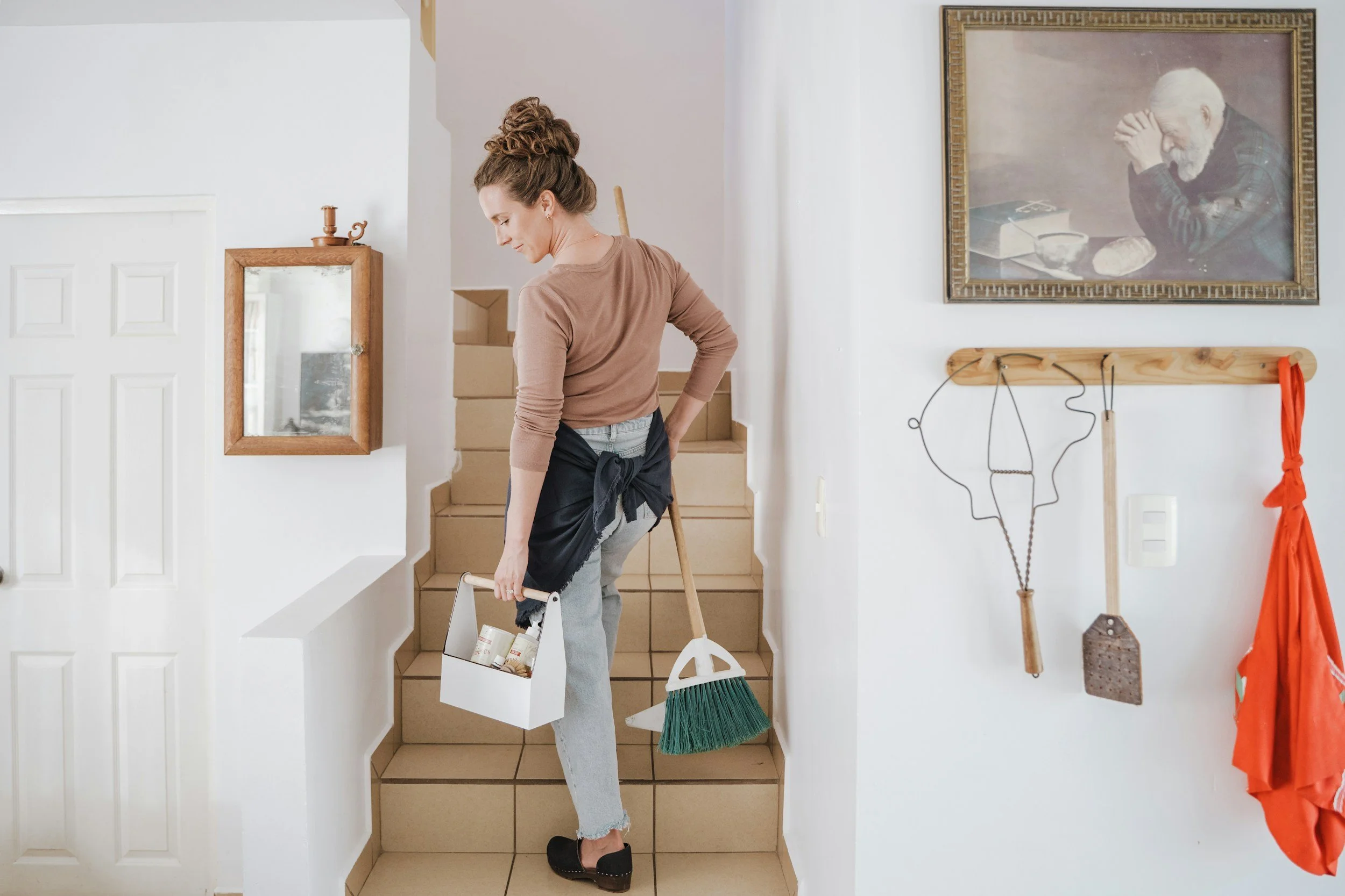 A woman with curly hair in a bun, wearing a brown long-sleeve shirt and jeans, is standing on stairs inside a house. She is holding a white basket with cleaning supplies and a mop. The interior has white walls, a framed mirror, and a painting of an elderly man with a beard. There are cleaning tools and an orange cloth hanging on the wall.