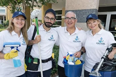 Four people in white shirts with an anchor logo, holding cleaning tools and supplies, standing outdoors smiling.