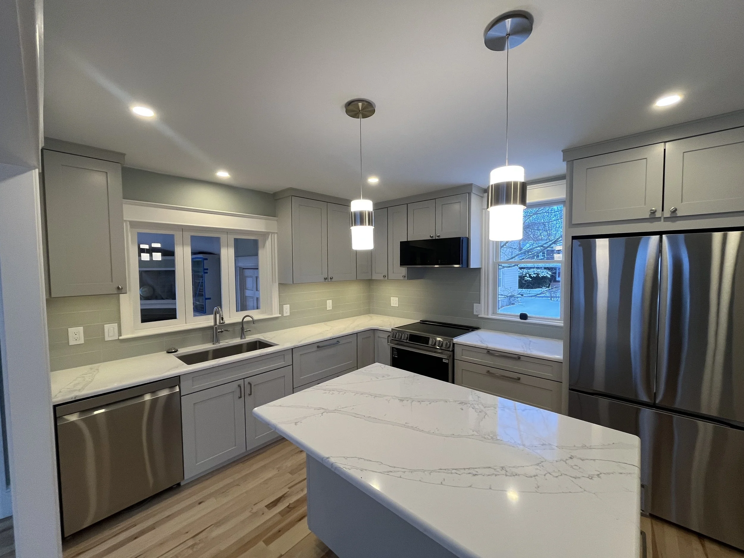 Modern kitchen with white cabinets, marble countertops, stainless steel appliances, pendant lights, and large windows showing snow outside.