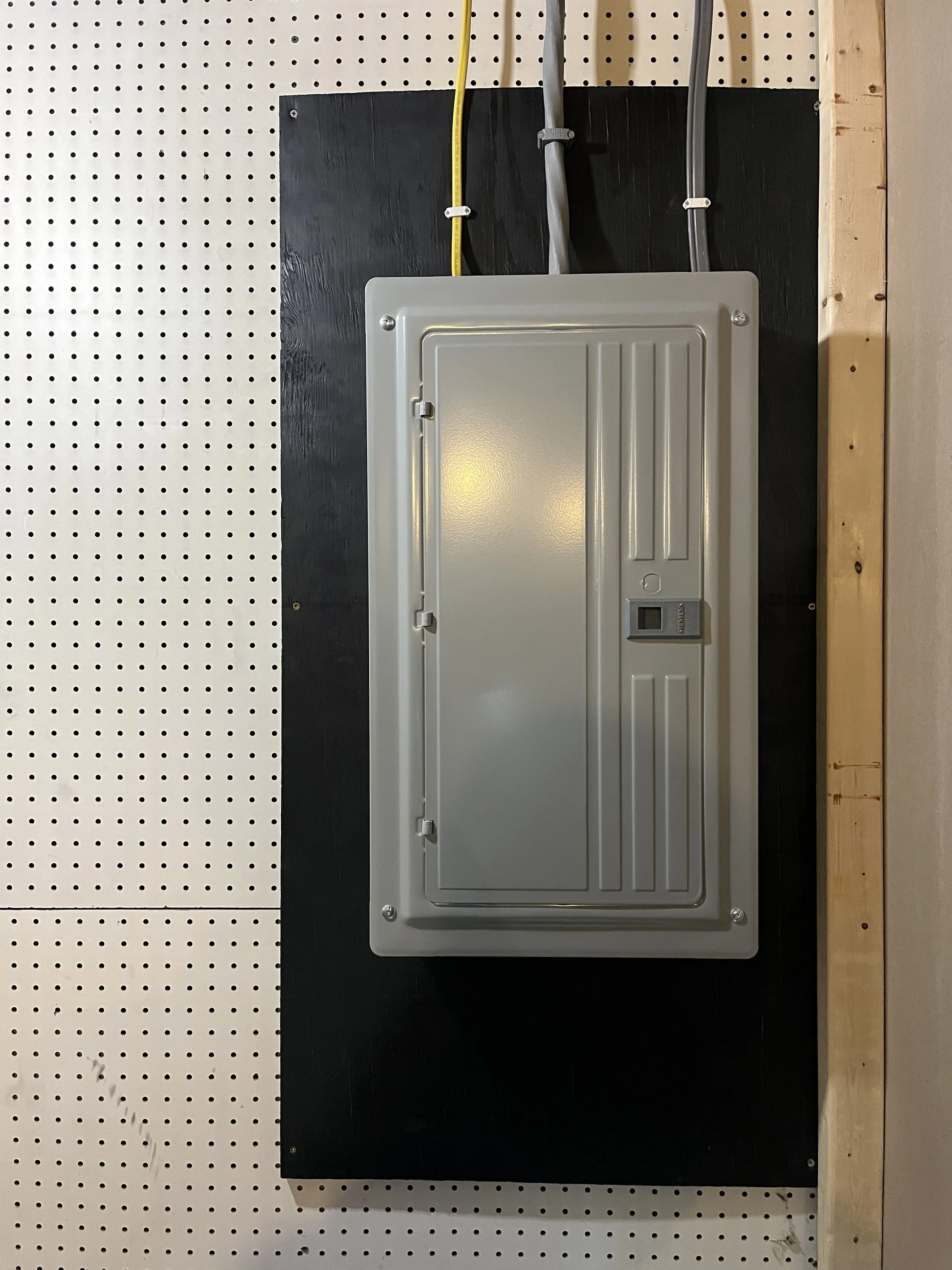 Electrical box mounted on a black board with electrical wires above it, set on a perforated white pegboard wall with a wooden frame.