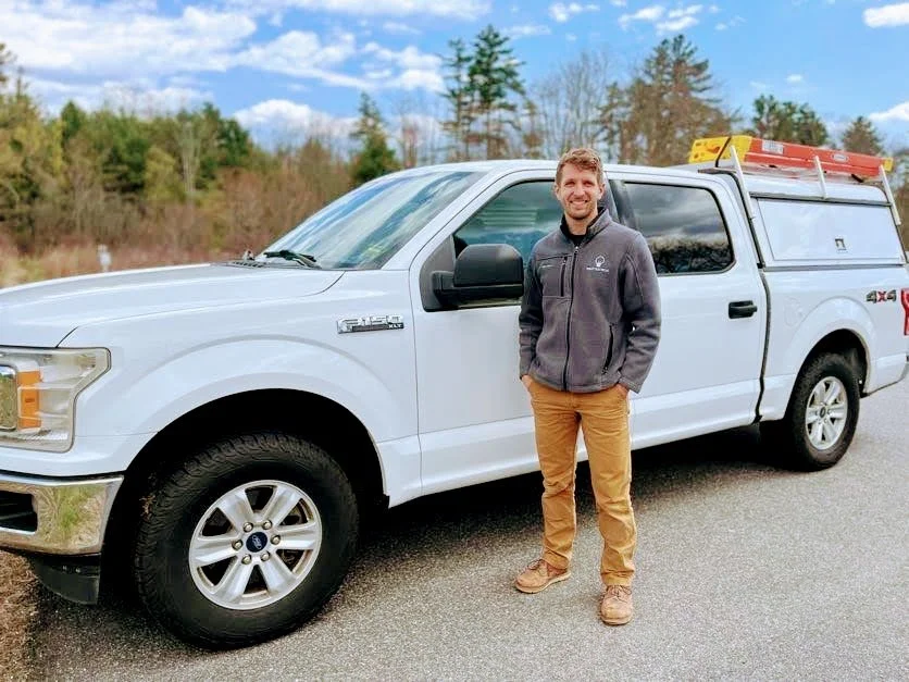 A friendly looking electrician standing next to a white work truck on a paved road with trees and a partly cloudy sky in the background.