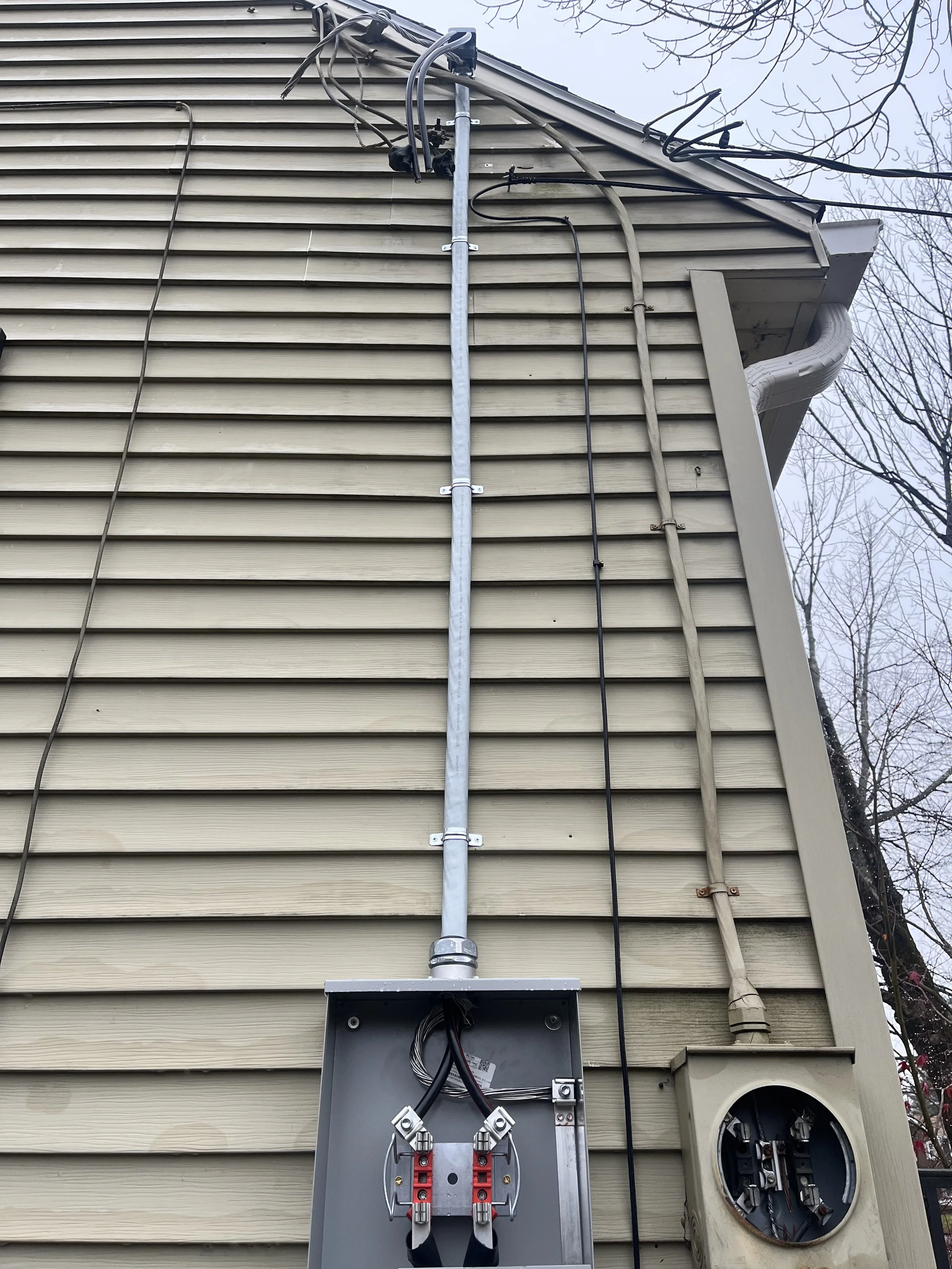 The exterior of a house with beige vinyl siding, showing electrical wiring and a metal conduit running vertically. There is an electrical box with wires inside, and a circular electric meter on the side of the house. The house has a roofline with a gutter and some leafless tree branches are visible against the sky.