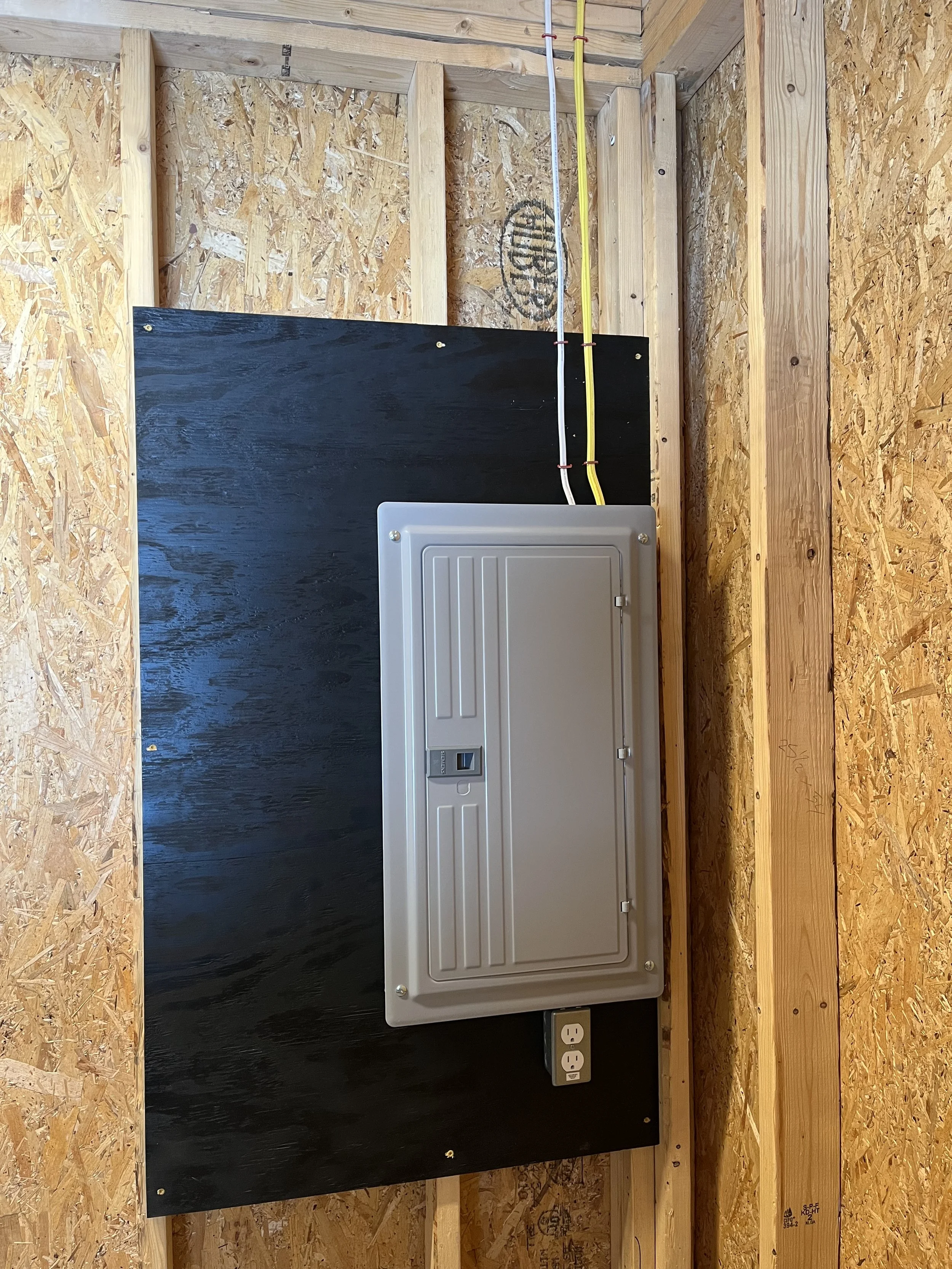 Construction wall with electrical box, wiring, and outlets mounted on a black panel, with wooden framing and OSB sheathing.