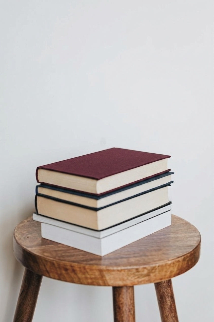 Four hardcover books stacked on a wooden stool against a plain wall.