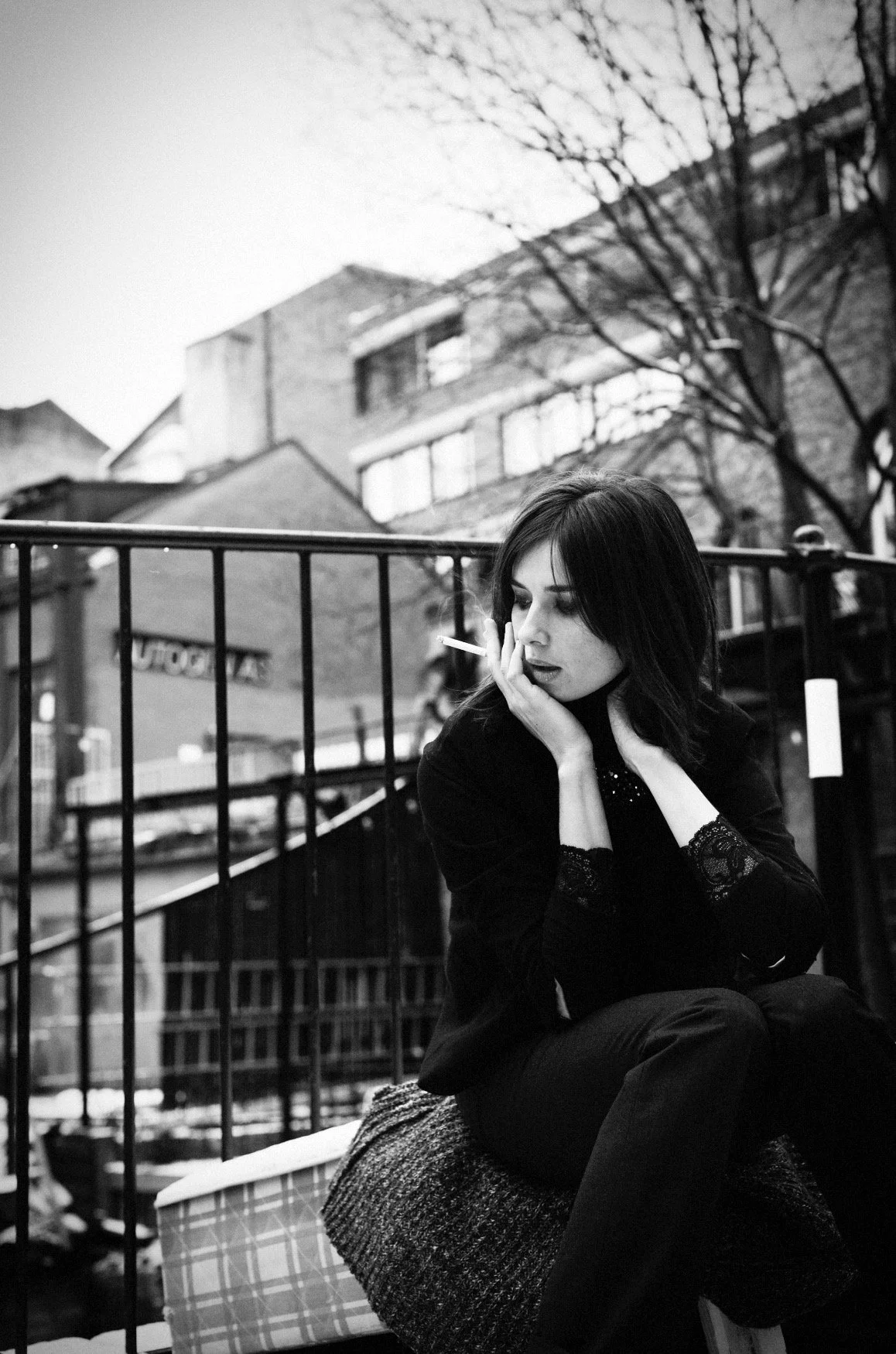 A black-and-white photo of a woman with dark hair sitting outdoors on a chair with a blanket. She appears to be contemplating, with one hand resting on her face. Behind her is a metal fence, and in the background are buildings, a tree, and a bicycle.