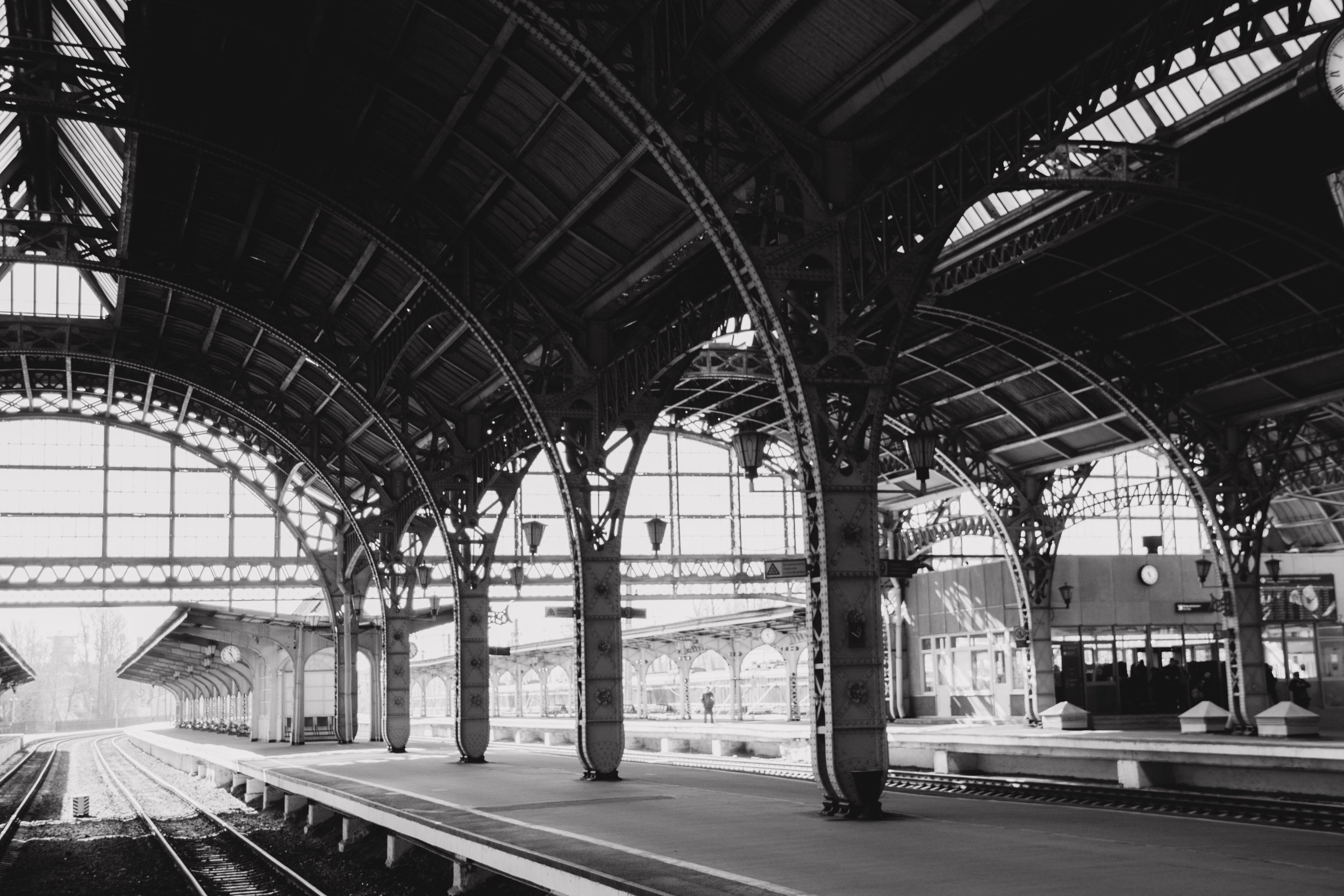 Interior view of a historic train station with arched metal and glass roof structure, train tracks, and platform. Black and white photo with a vintage feel.