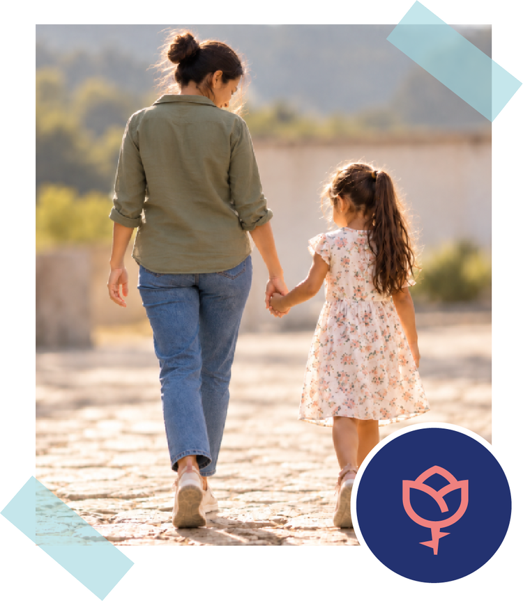 A woman and a young girl walk hand in hand down a sunlit path, seen from behind