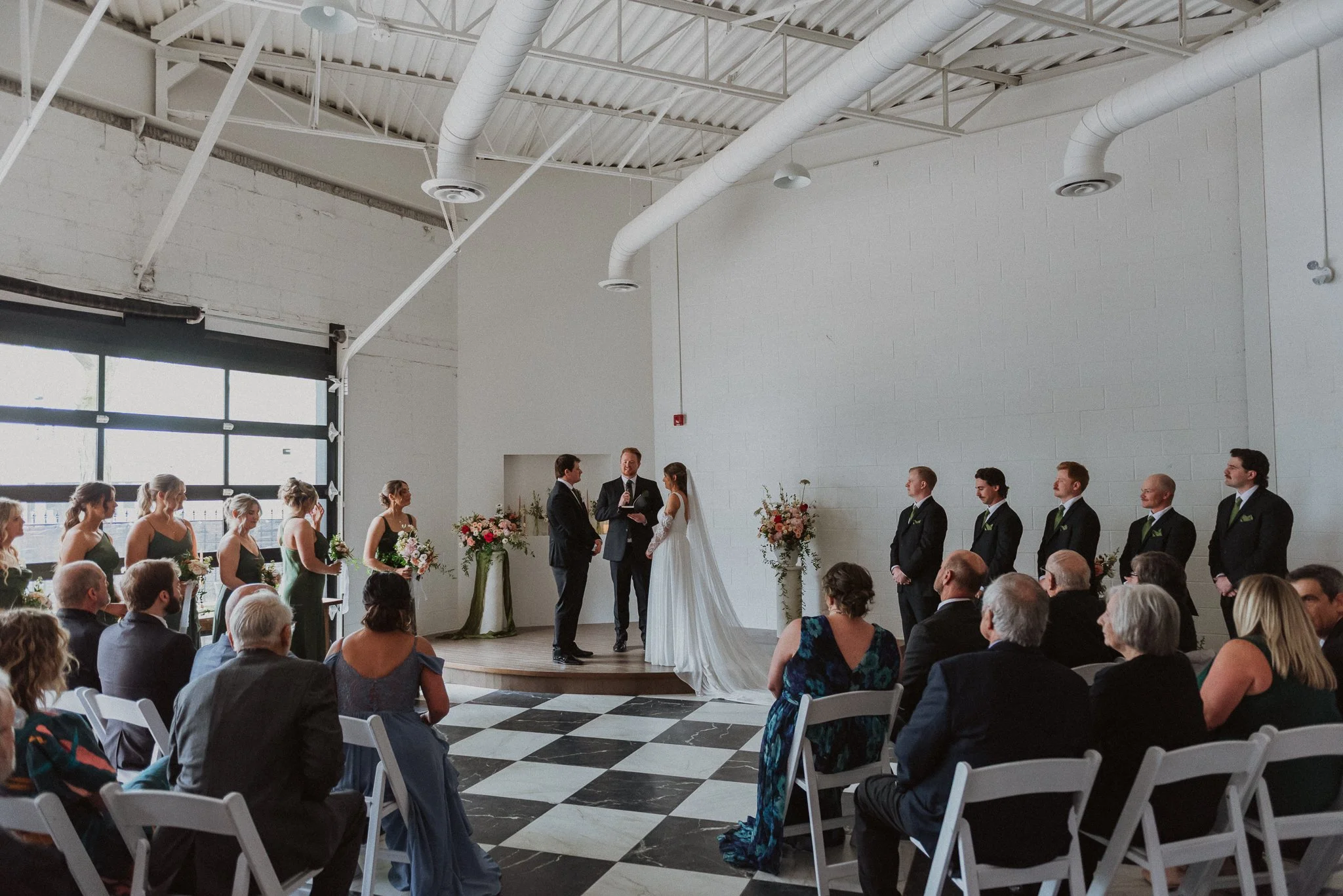 Indoor wedding ceremony in a wedding venue with high ceilings and architectural details and a unique checkerboard dance floor, large windows, and industrial ceiling with white ductwork.