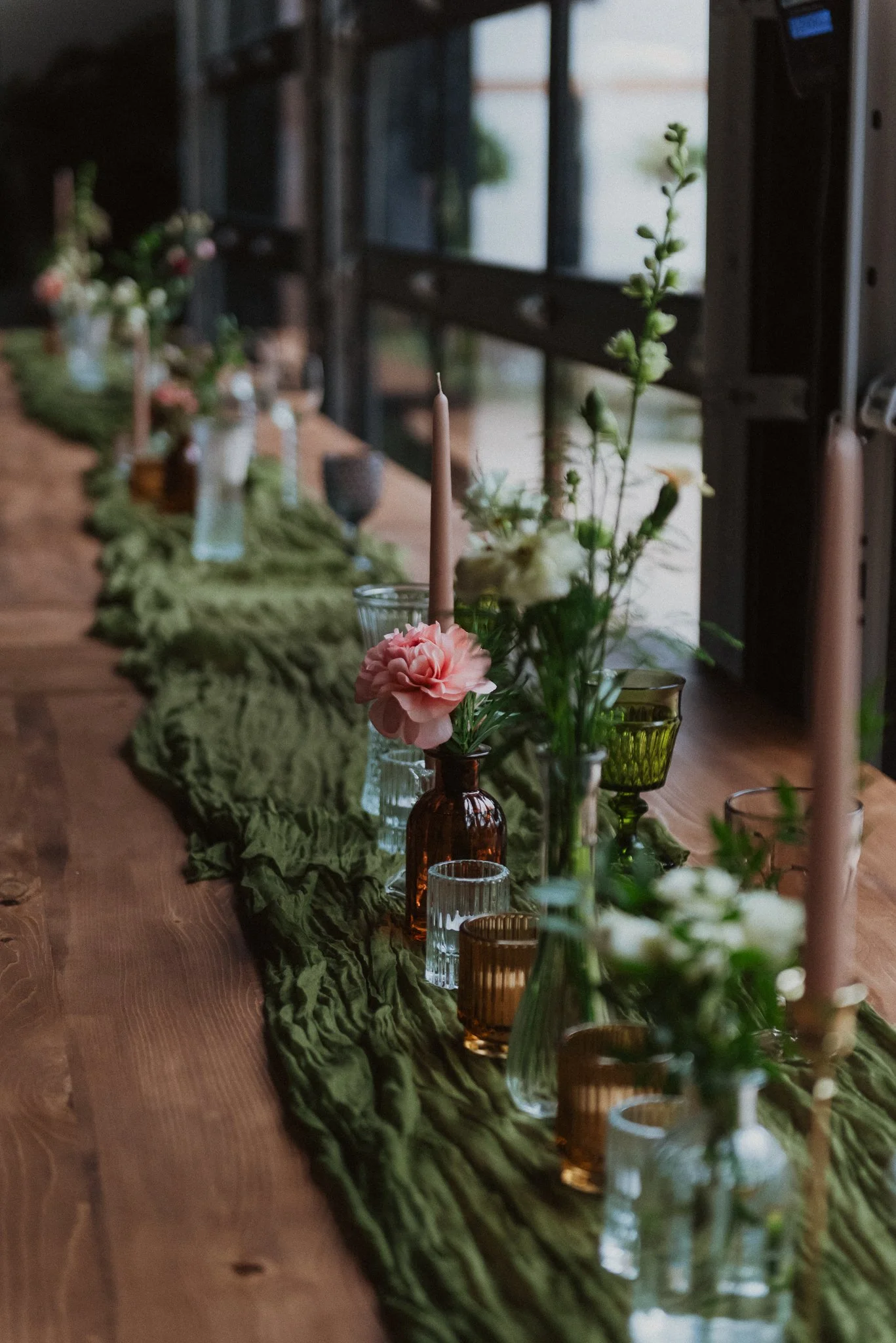 A long wooden table decorated with a green fabric table runner, candles, and various flower arrangements in glass jars and bottles inside a rustic, industrial-style building with large windows.