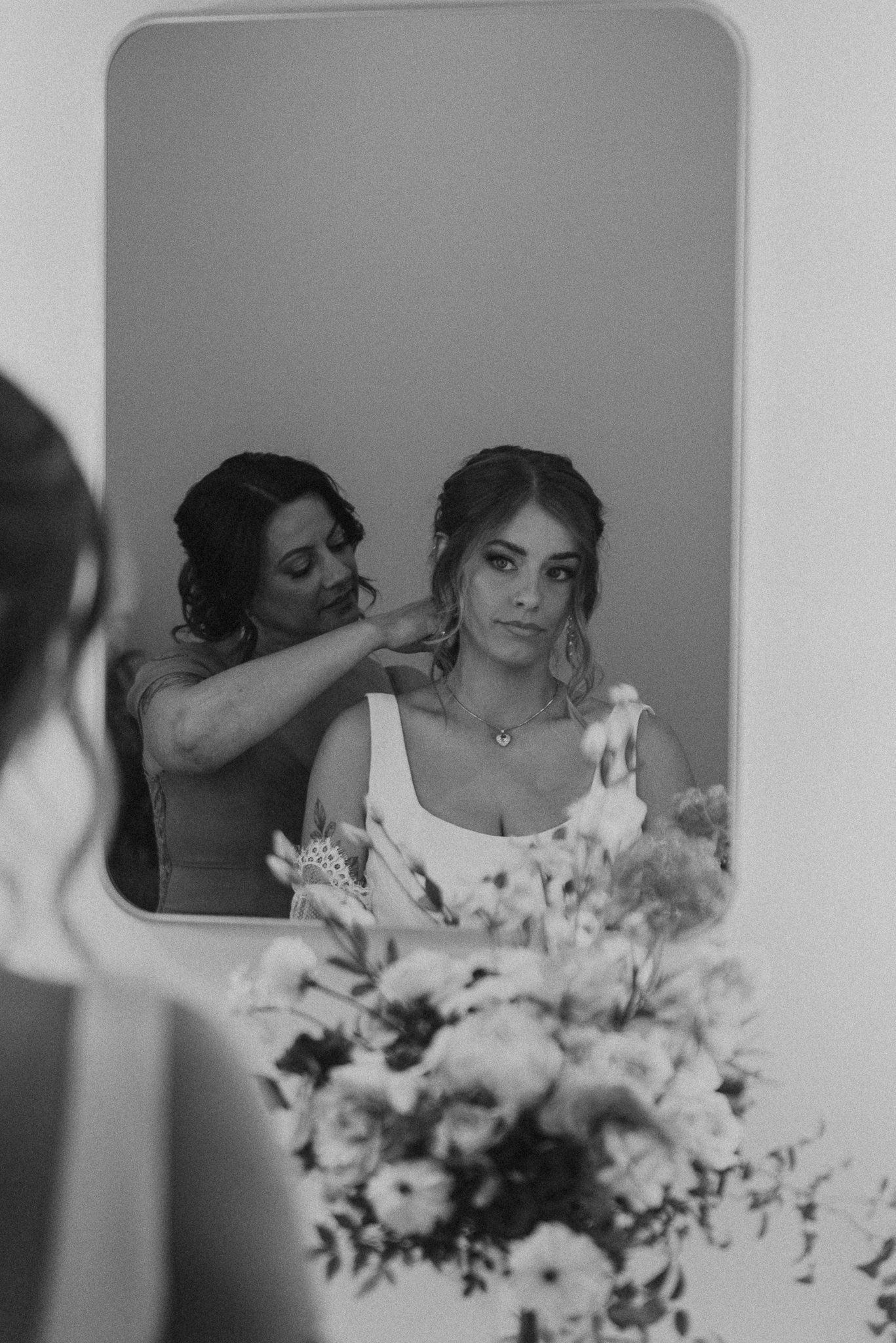 Black and white photo of a woman getting ready, seen through a mirror, with flowers in the foreground.