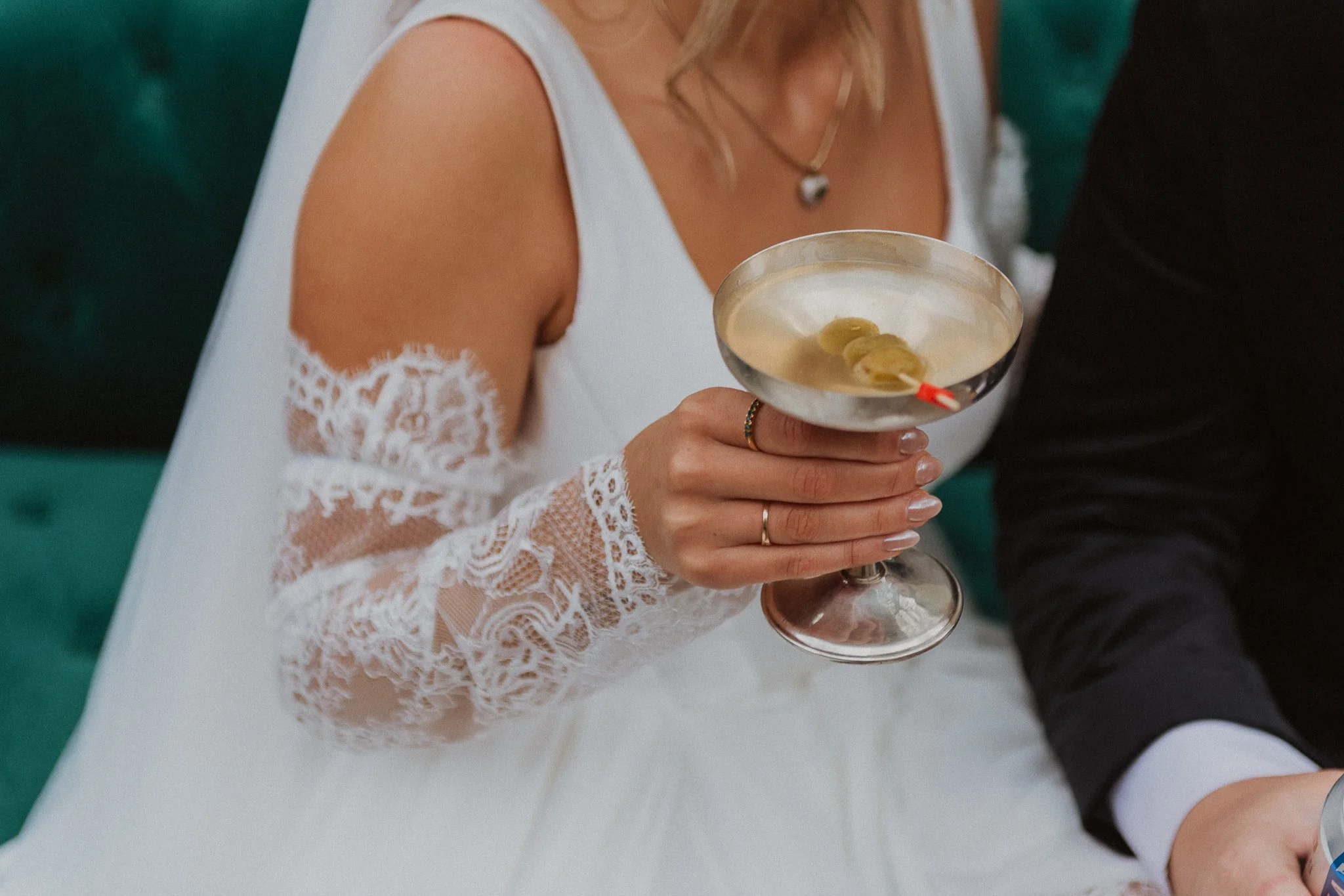 A woman in a white dress with lace sleeves holds a cocktail glass with olives inside. She wears a necklace and has blonde hair.