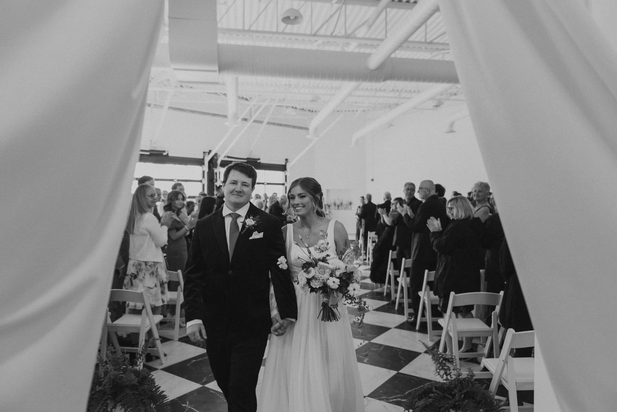 A newly married couple is walking down the aisle during a wedding ceremony, holding hands and smiling, surrounded by seated guests in a unique non-traditional wedding venue