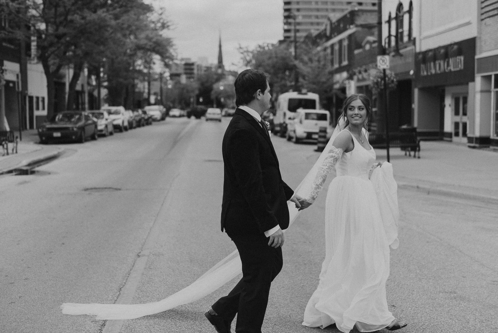 A newlywed couple holding hands while walking in downtown Sarnia on their wedding day, with cars parked along the side and buildings in the background.