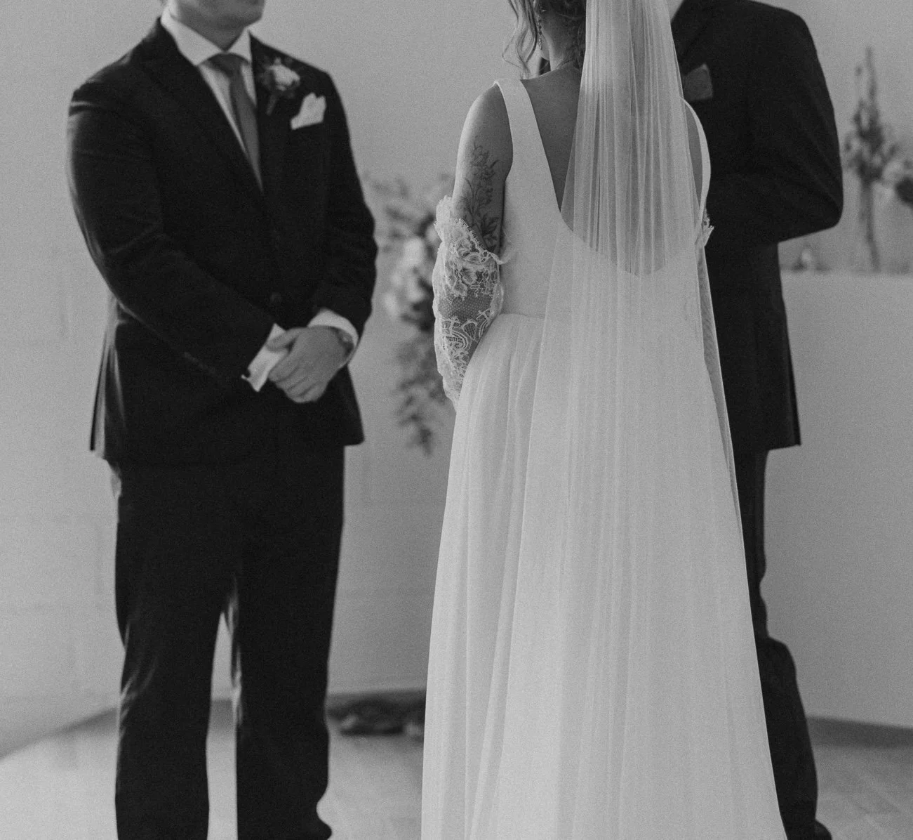 A black-and-white photo of a wedding ceremony showing a bride and groom standing facing each other, with an officiant present. The bride wears a long white dress and a veil, and the groom wears a dark suit with a boutonniere. The bride's dress has lace sleeves, and she has visible tattoos on her arms.