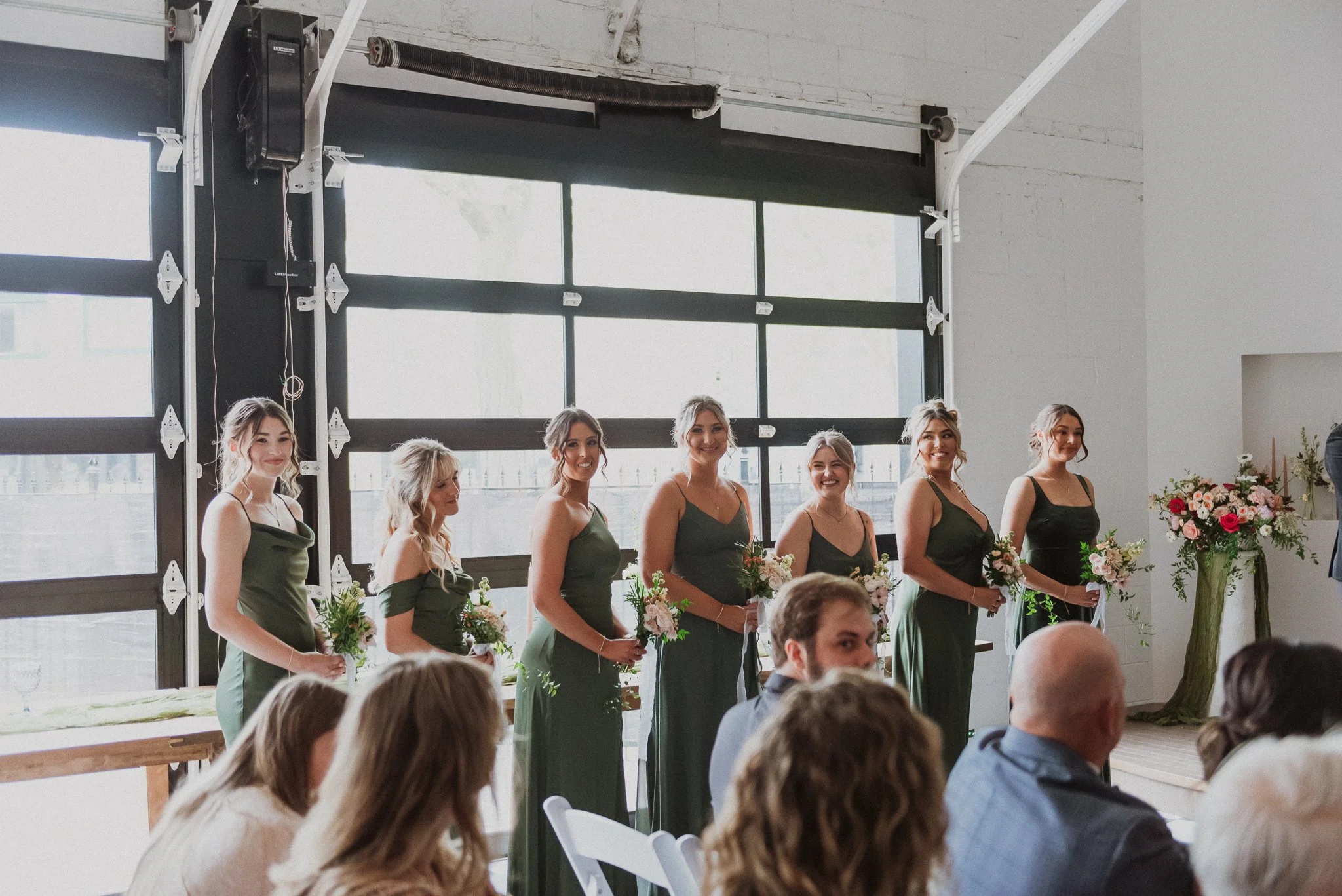 A group of bridesmaids standing in a row during a wedding ceremony, holding bouquets of flowers, inside a modern industrial-style venue with large windows.