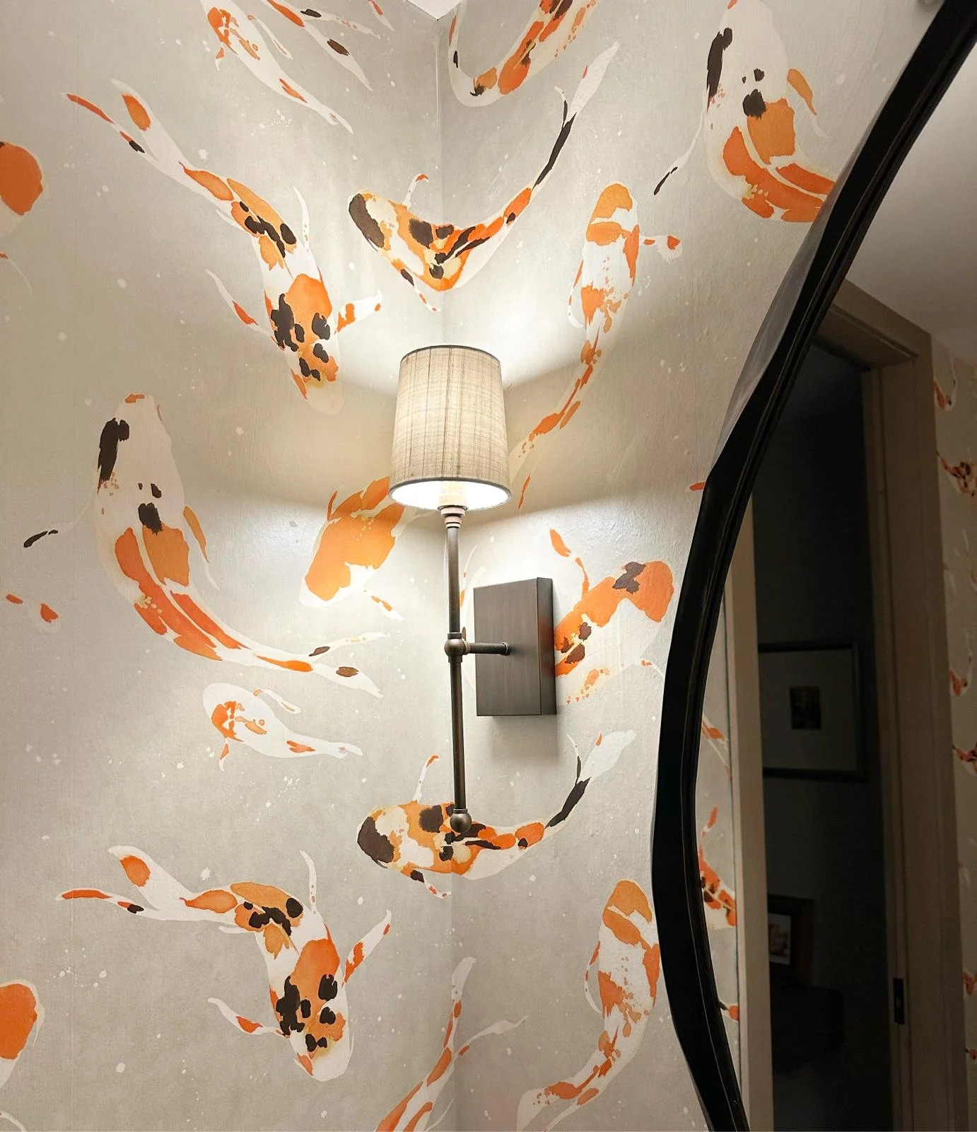 A wall with wallpaper of orange, white, and black koi fish, illuminated by a wall-mounted lamp with a beige lampshade. A black-framed mirror is partially visible on the right side.