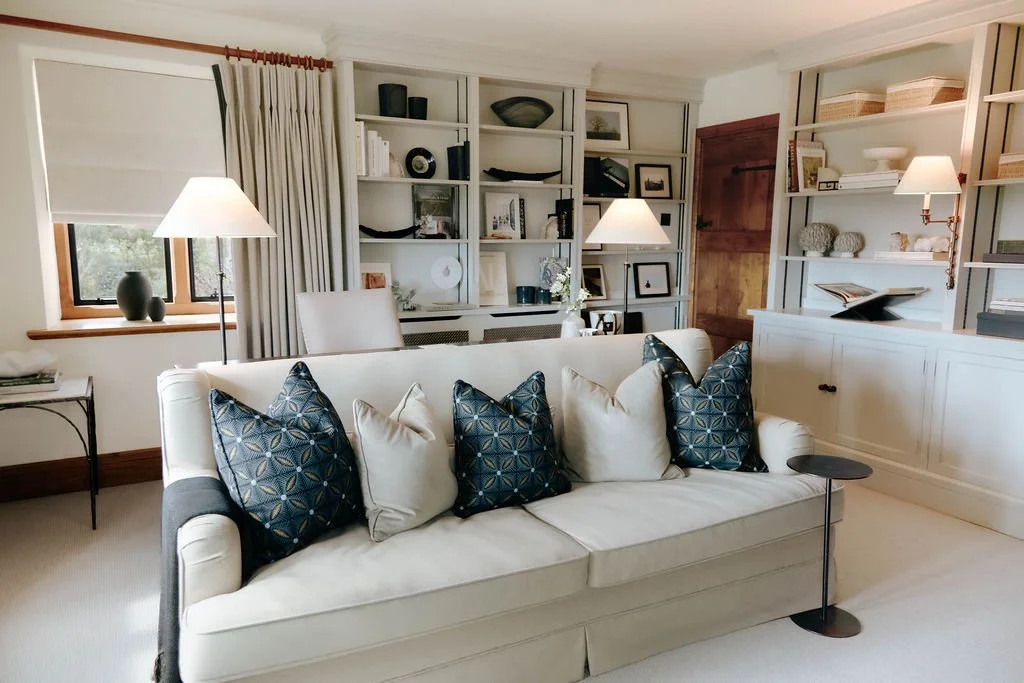 Living room with a white sofa adorned with blue and white pillows, a window with beige curtains, and built-in white bookshelves filled with decorative items, framed pictures, and books. There are two floor lamps providing lighting.