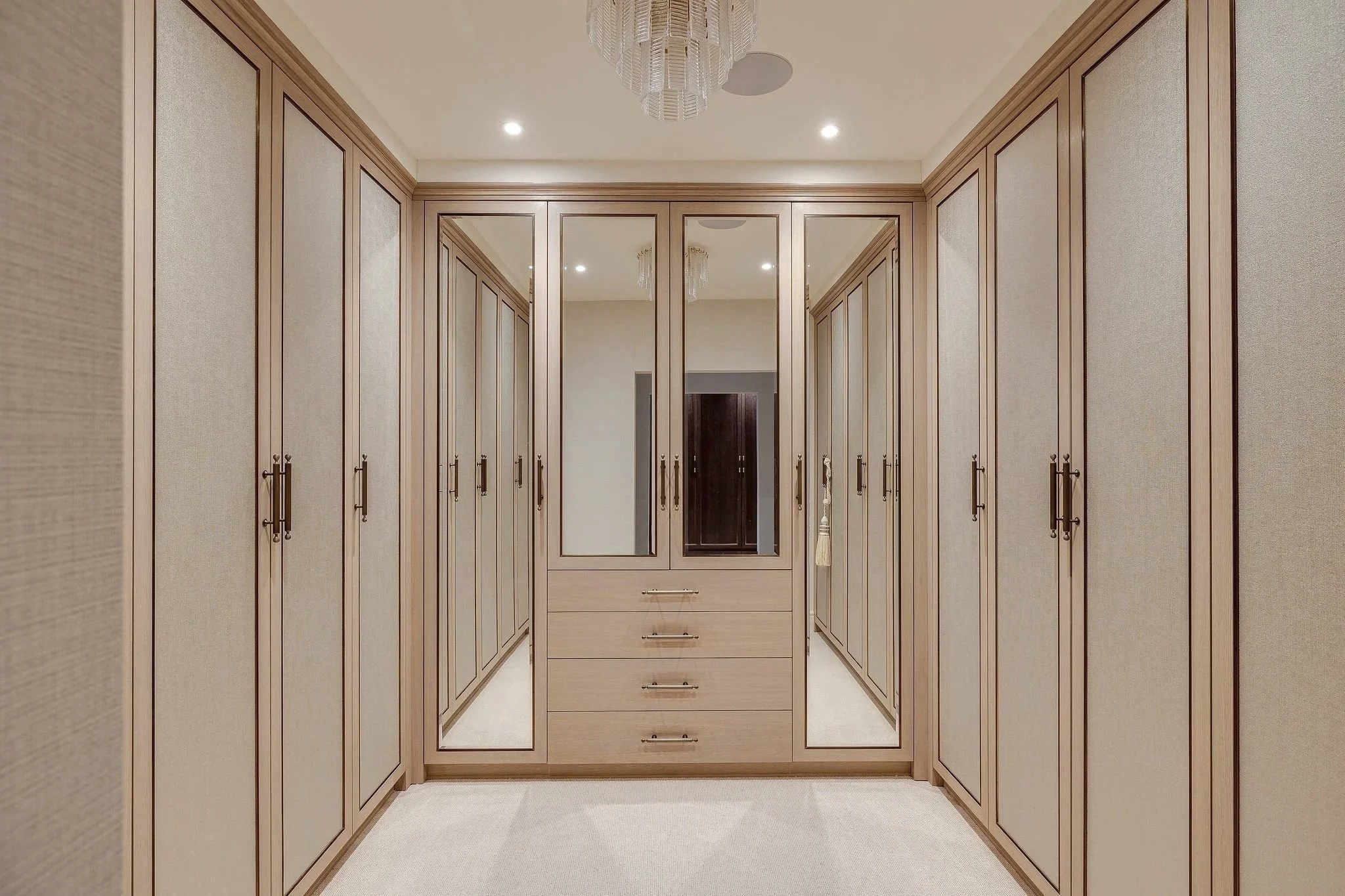 A walk-in wardrobes with built-in wooden cabinets and drawers, beige textured wallpaper, and a cream carpet. A chandelier hangs from the ceiling.
