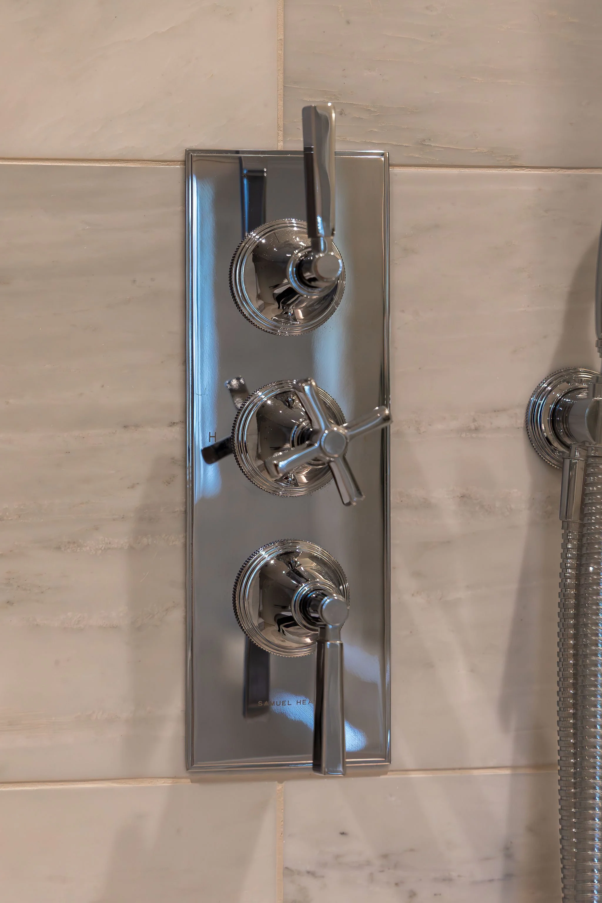 Close-up view of a modern shower valve with three chrome knobs mounted on a beige tiled wall.