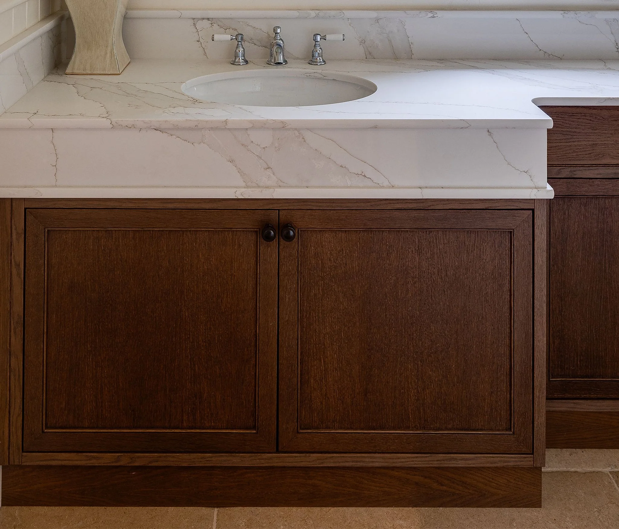 A bathroom vanity with a white marble countertop and a dark wooden cabinet. The countertop has a round, inset sink with a chrome faucet and two handles. There is a decorative wooden object on the left side of the countertop.