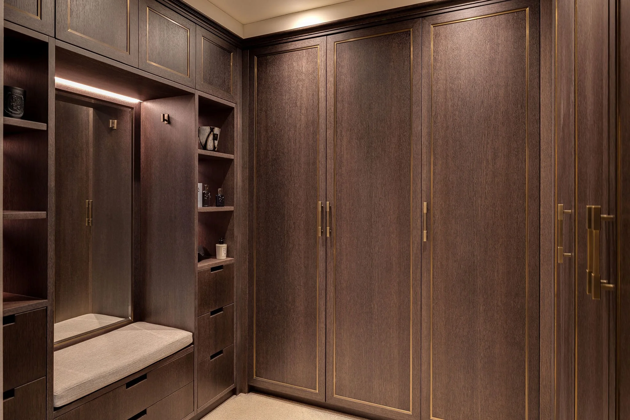 A closet with dark wood paneling, gold accents, a built-in bench with a cushion, and shelves with decorative items.