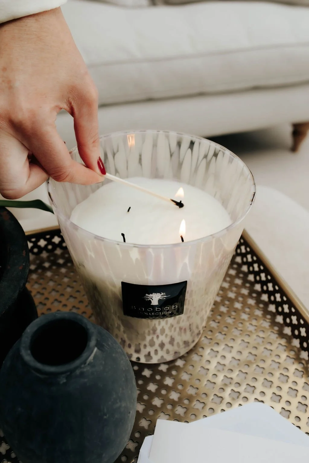 A person with red painted nails lighting the wick of a large white scented candle with a matchstick. The candle is in a clear glass holder on a decorative tray, with a dark vase nearby.