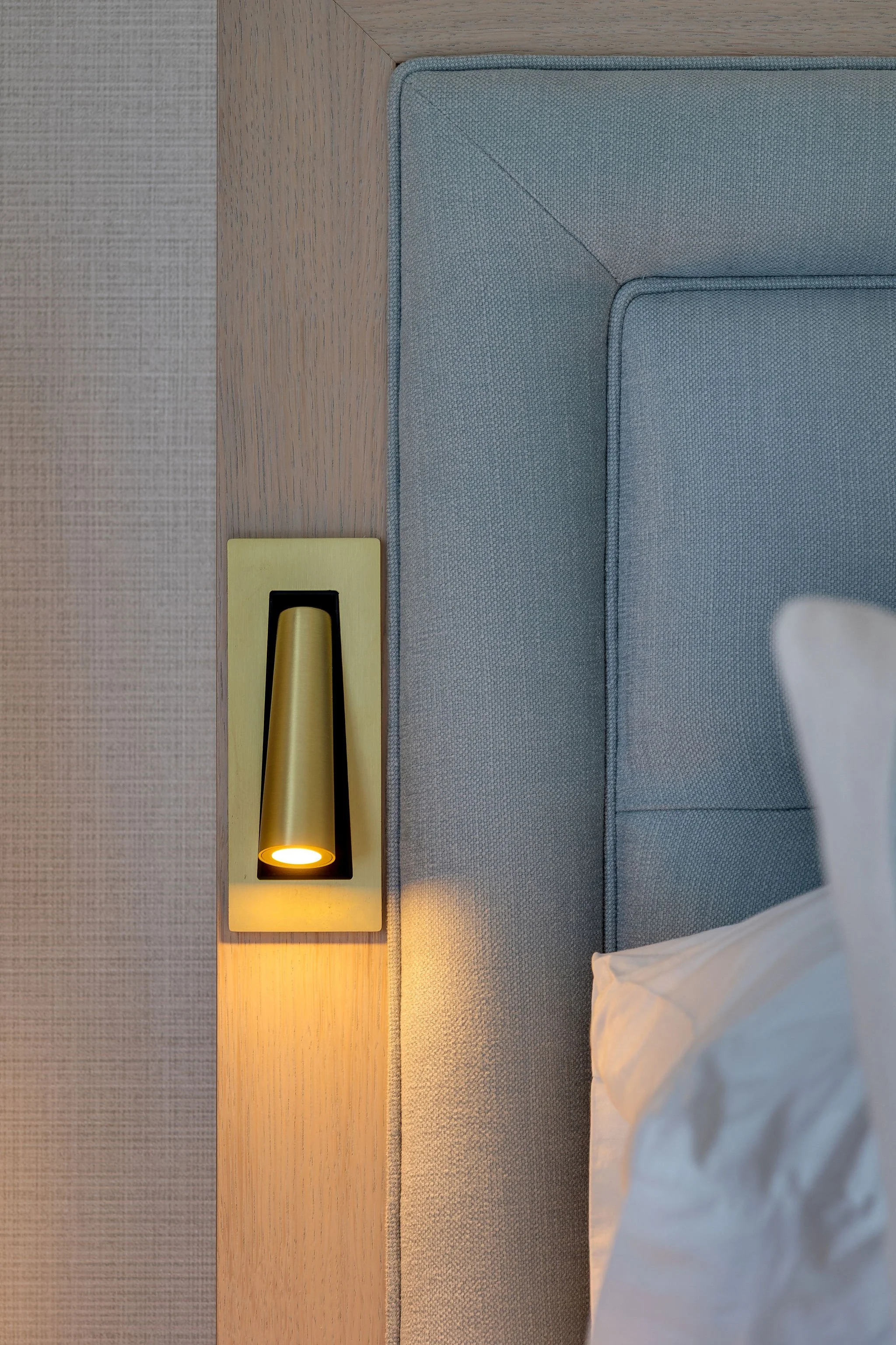 Close-up of a modern bed with grey upholstered headboard, wooden side table with yellow wall-mounted reading light, and a white pillow in a hotel room.