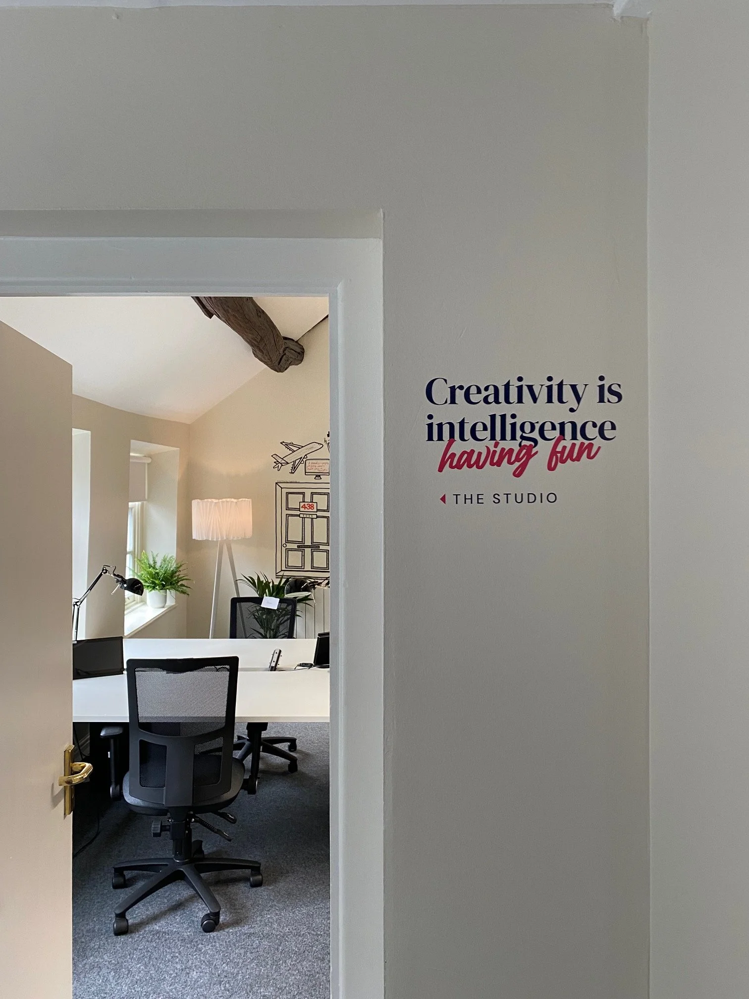 Office space seen through an open door with a white desk, black office chair, plants, and wall decorations.