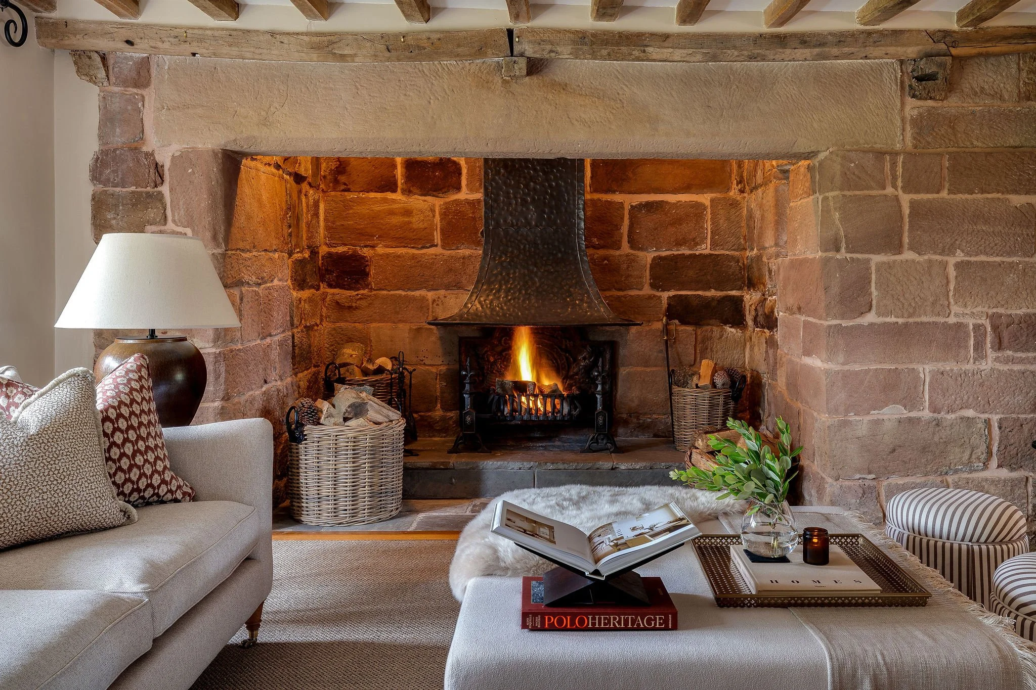 Living room with a fireplace, beige sofa, patterned pillows, wicker baskets, a lamp, a coffee table with books and a plant, and a blanket draped on the sofa.