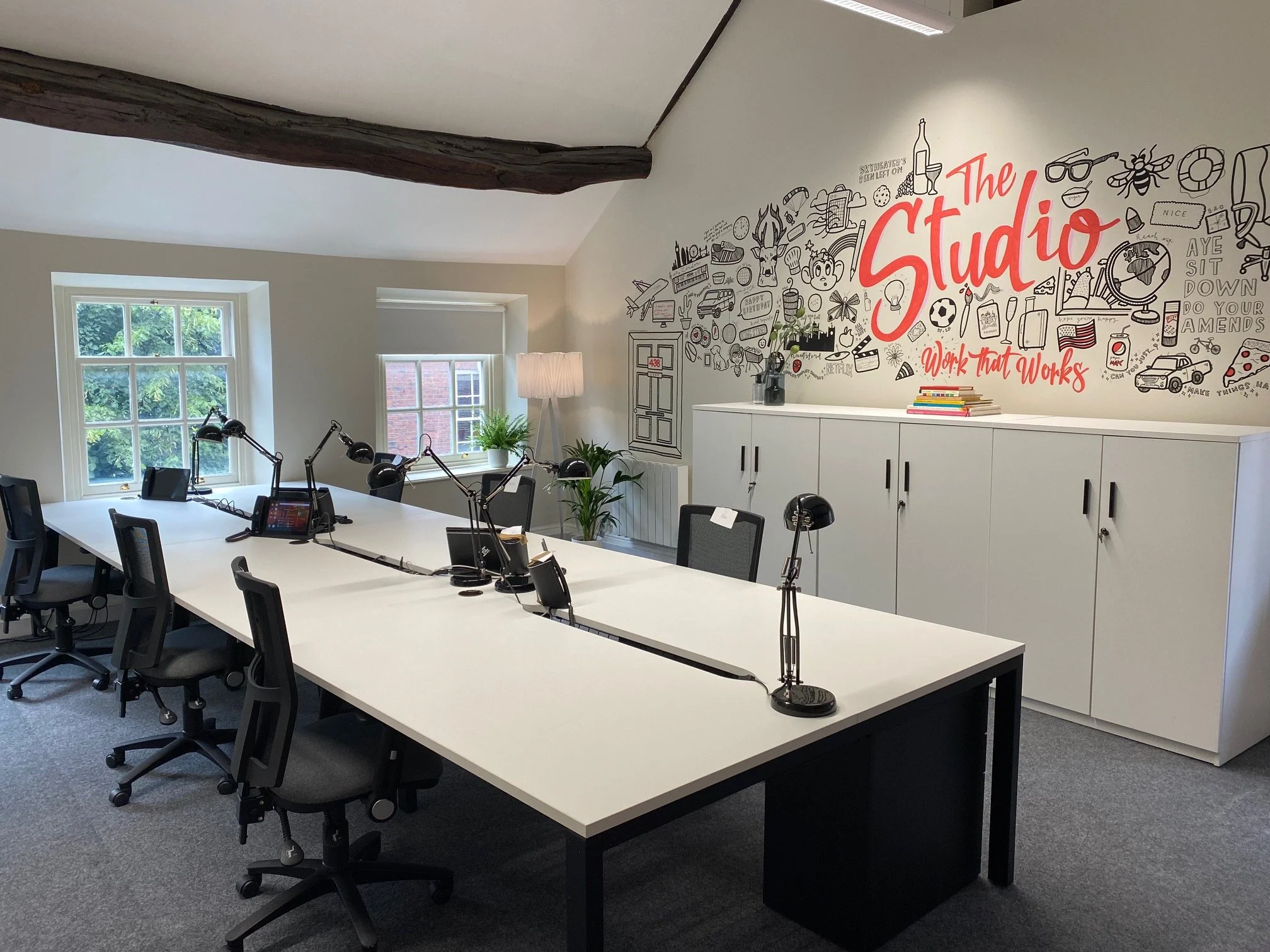 Modern office conference room with white tables and black chairs, two windows showing green foliage outside, and a feature wall with a large mural reading 'The Studio' surrounded by various doodles and icons.