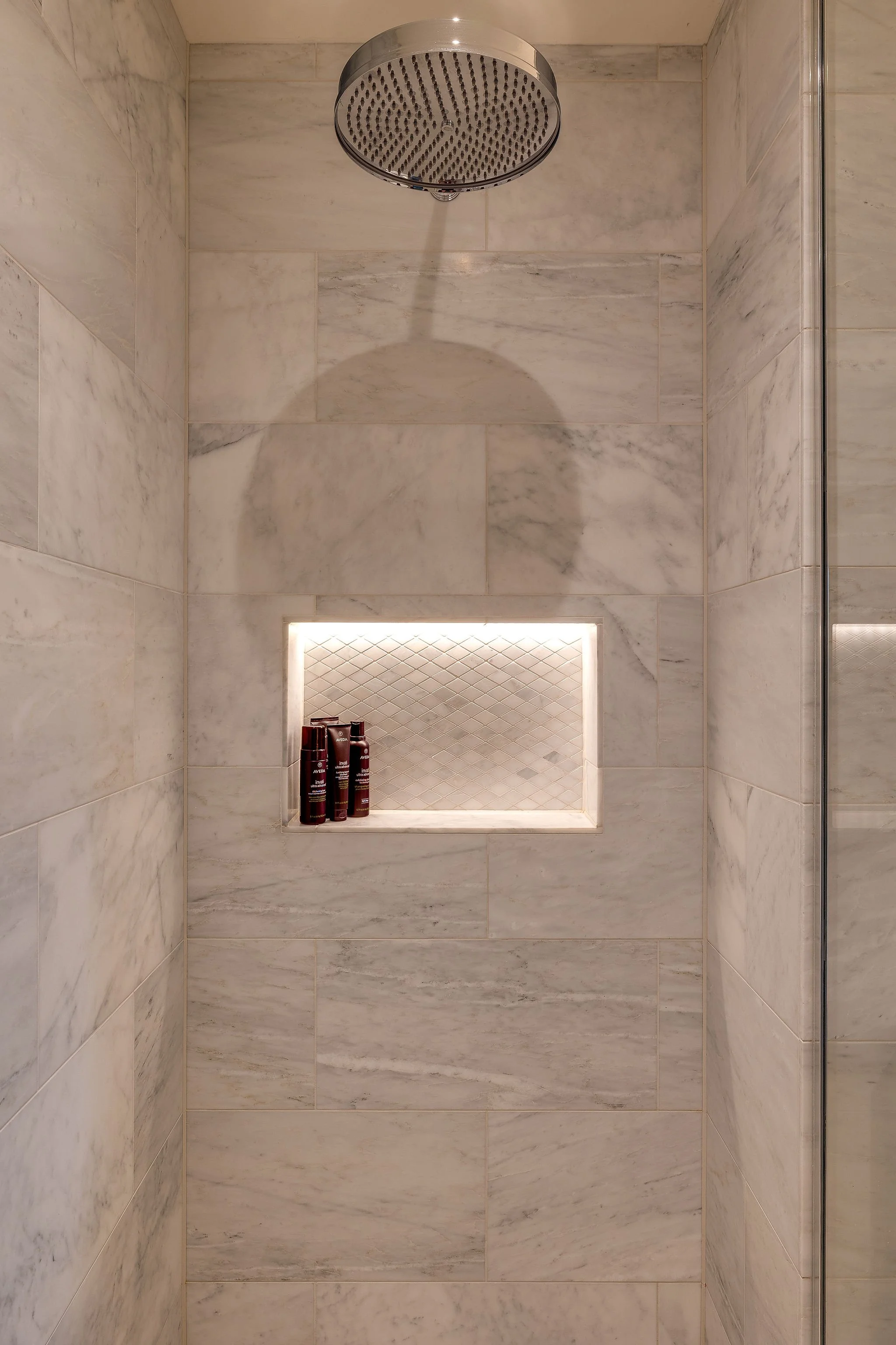 Shower area with a rainfall showerhead, large marble tiles on walls, a built-in shelf with three bottles, and a niche with a backlit design.