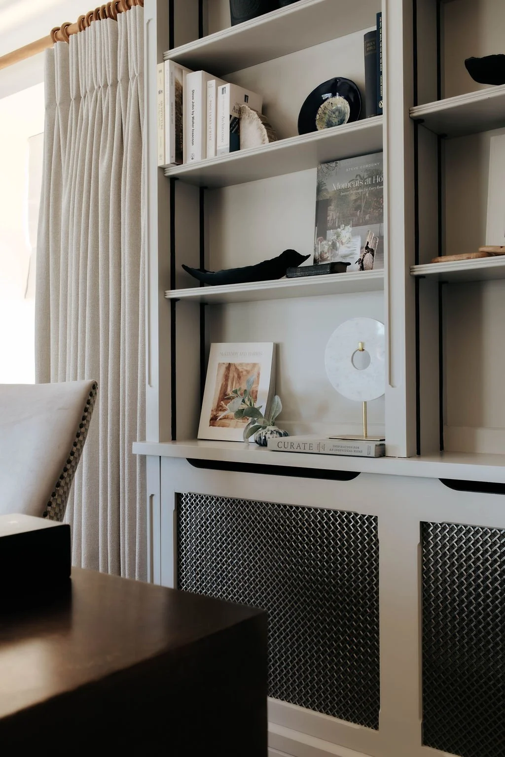 A white bookshelf with various decorative items, books, and artwork, with beige curtains on the side.